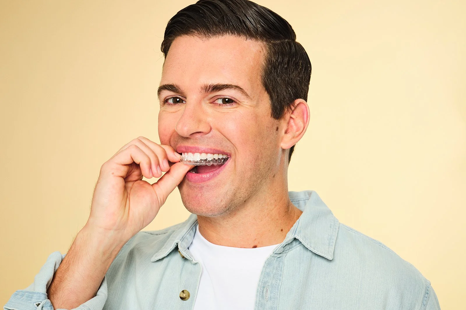 Man with dark hair smiling and holding a dental aligner near his teeth against a yellow background.