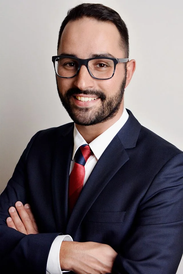 A professional man with dark hair, glasses, a beard, and a mustache, wearing a dark suit, white shirt, and red tie, smiling with arms crossed against a plain background.