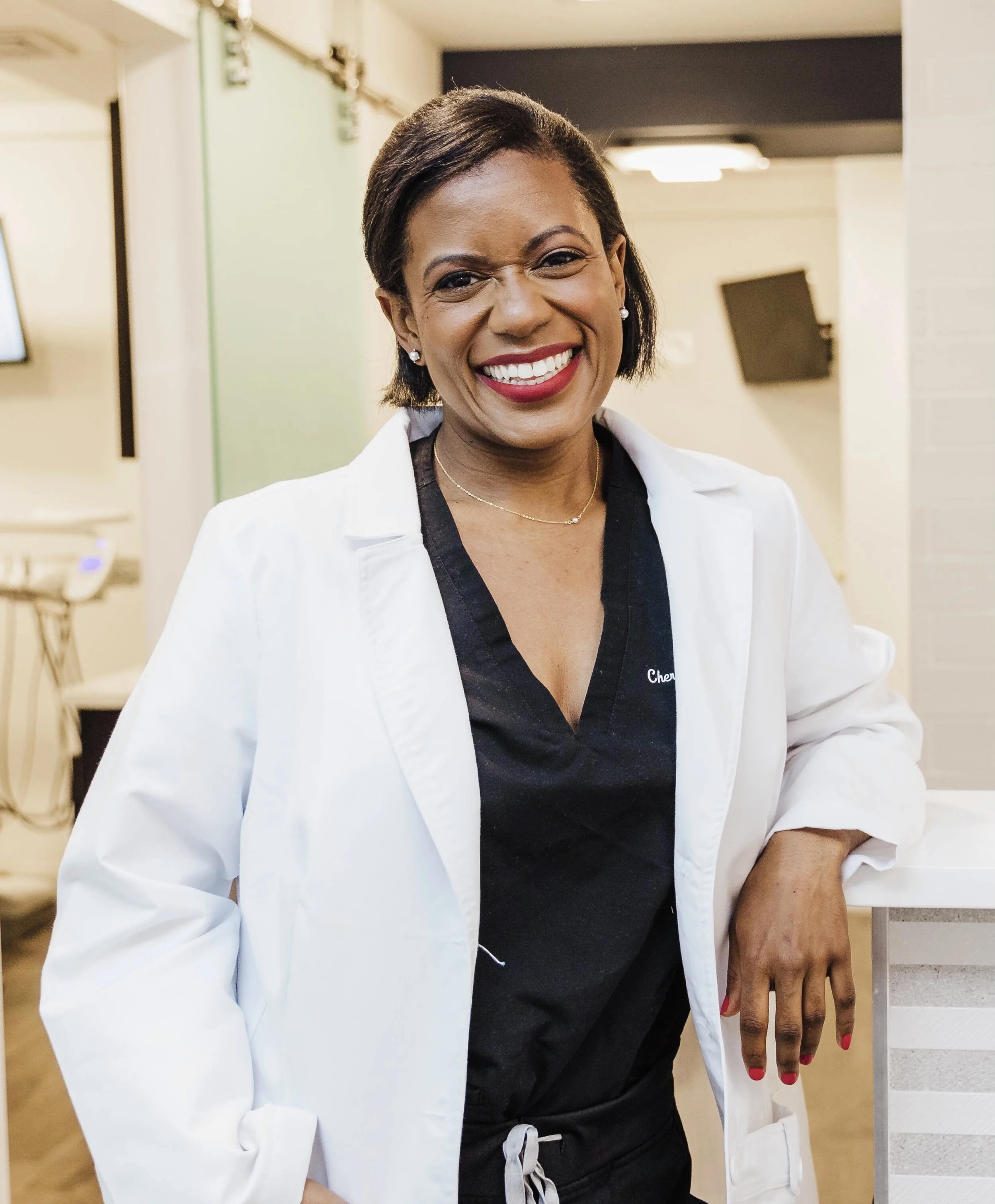 A smiling African American woman wearing a white medical coat and black scrubs, standing in a clinical or medical setting.