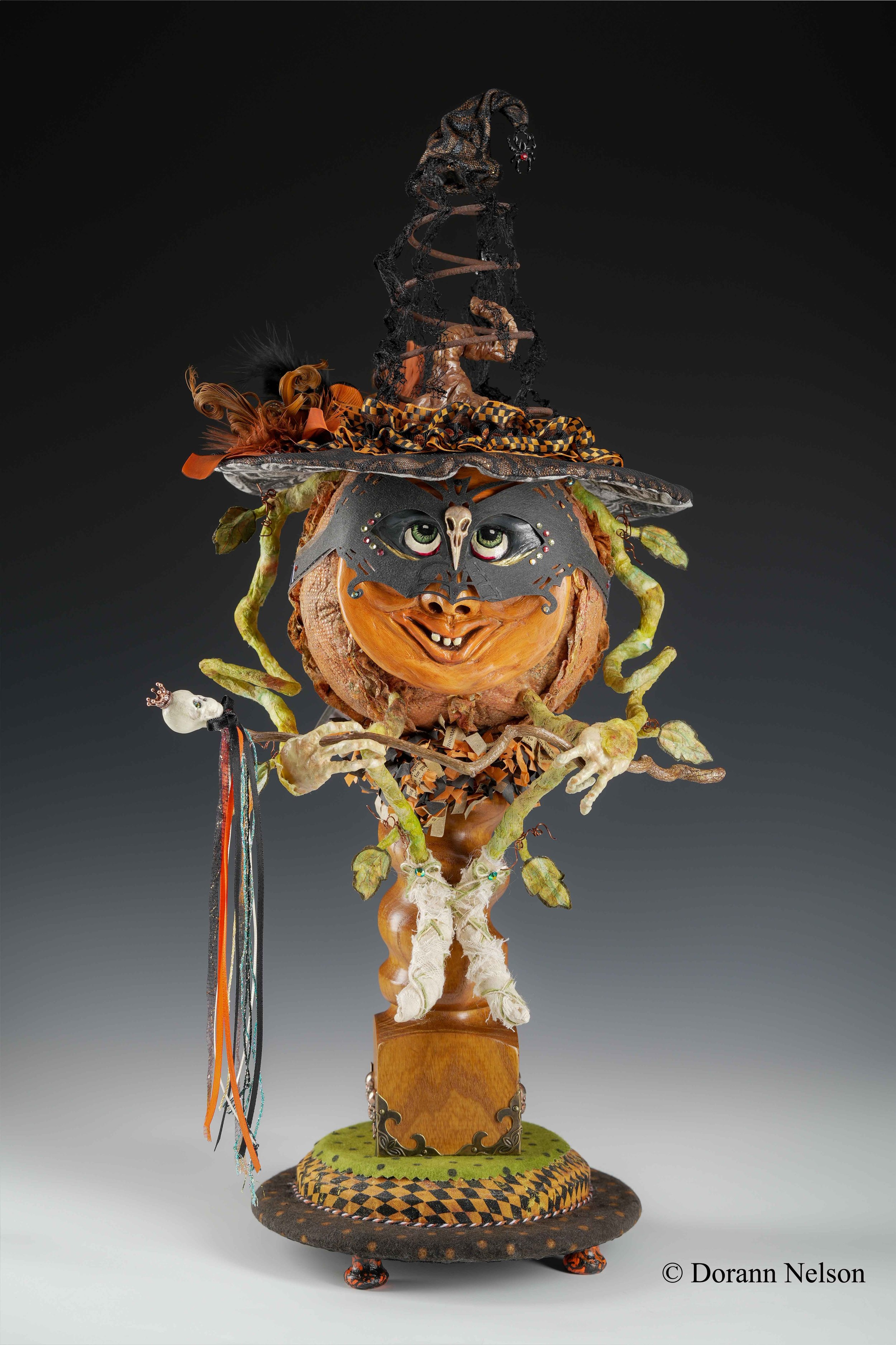 A whimsical mixed-media sculpture of a witch with a pumpkin head, wearing a pointy hat, adorned with various decorations, and holding a wand, standing on a round base.