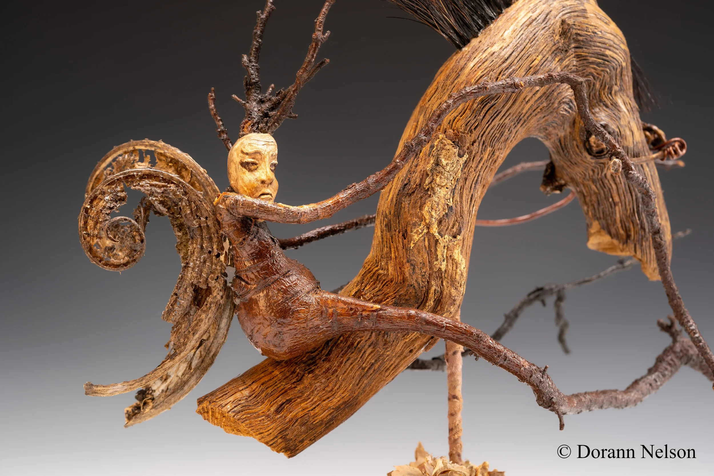 A sculpture of a humanoid figure with a face and arms, integrated into a twisted piece of wood and branches, with a natural, earthy appearance.