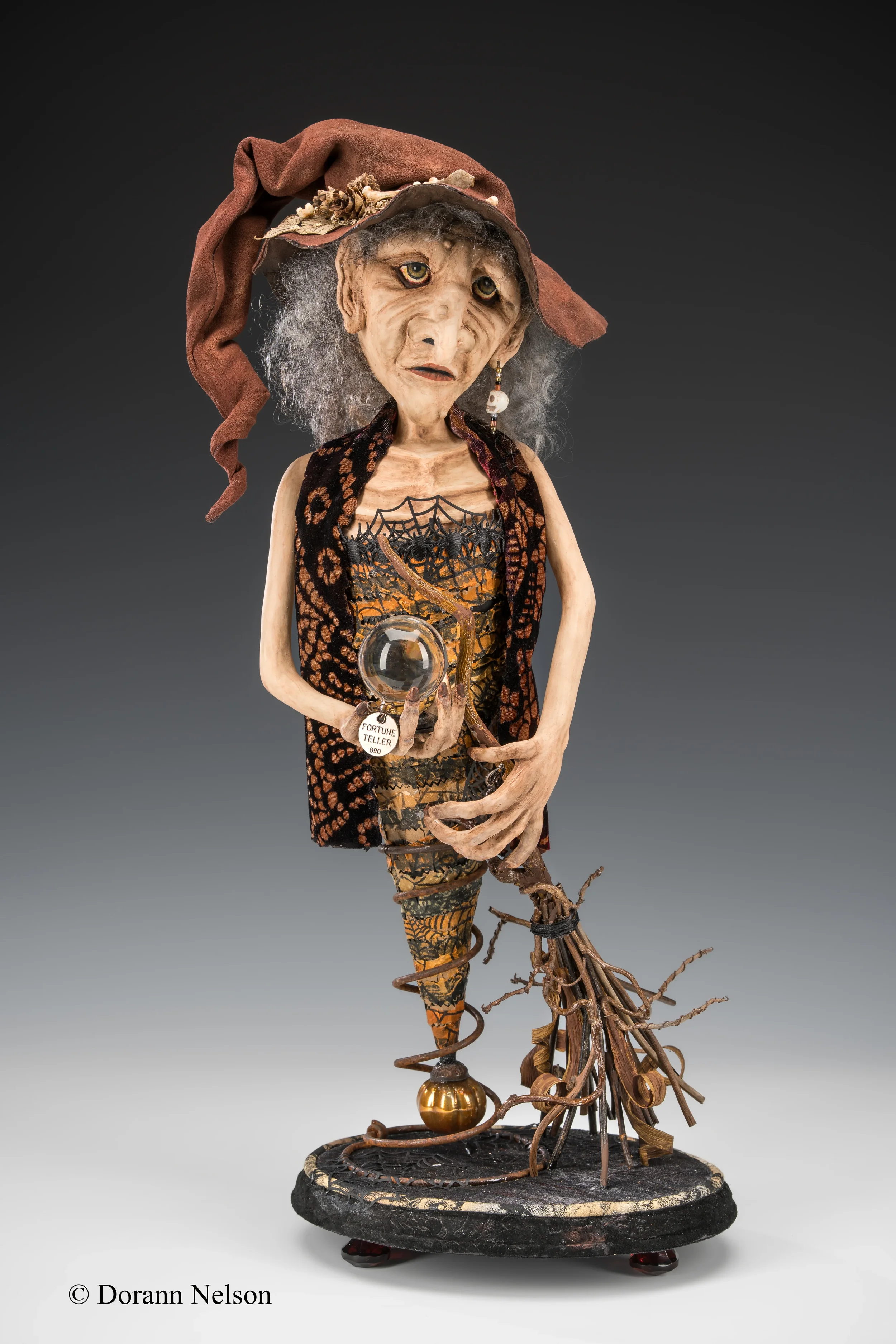 A whimsical sculpture of an elderly woman dressed as a fortune teller, wearing a witch's hat, holding a crystal ball, with a broom, dressed in patterned clothing, and with exaggerated facial features and gray curly hair.