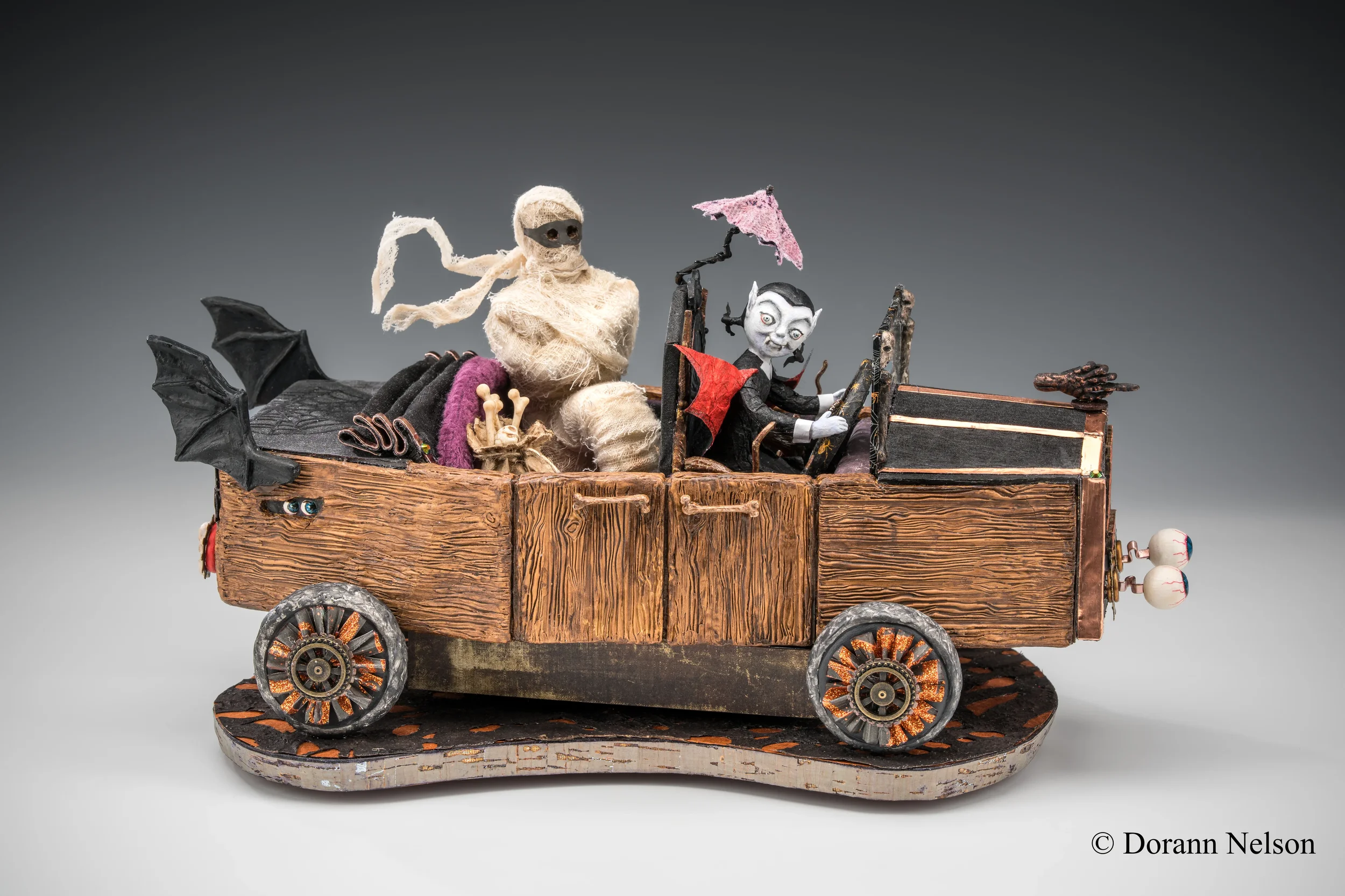 A handcrafted diorama featuring puppets and miniature objects inside a wooden vehicle, with characters resembling a mummy and a vampire, along with decorative elements like an umbrella and bats.
