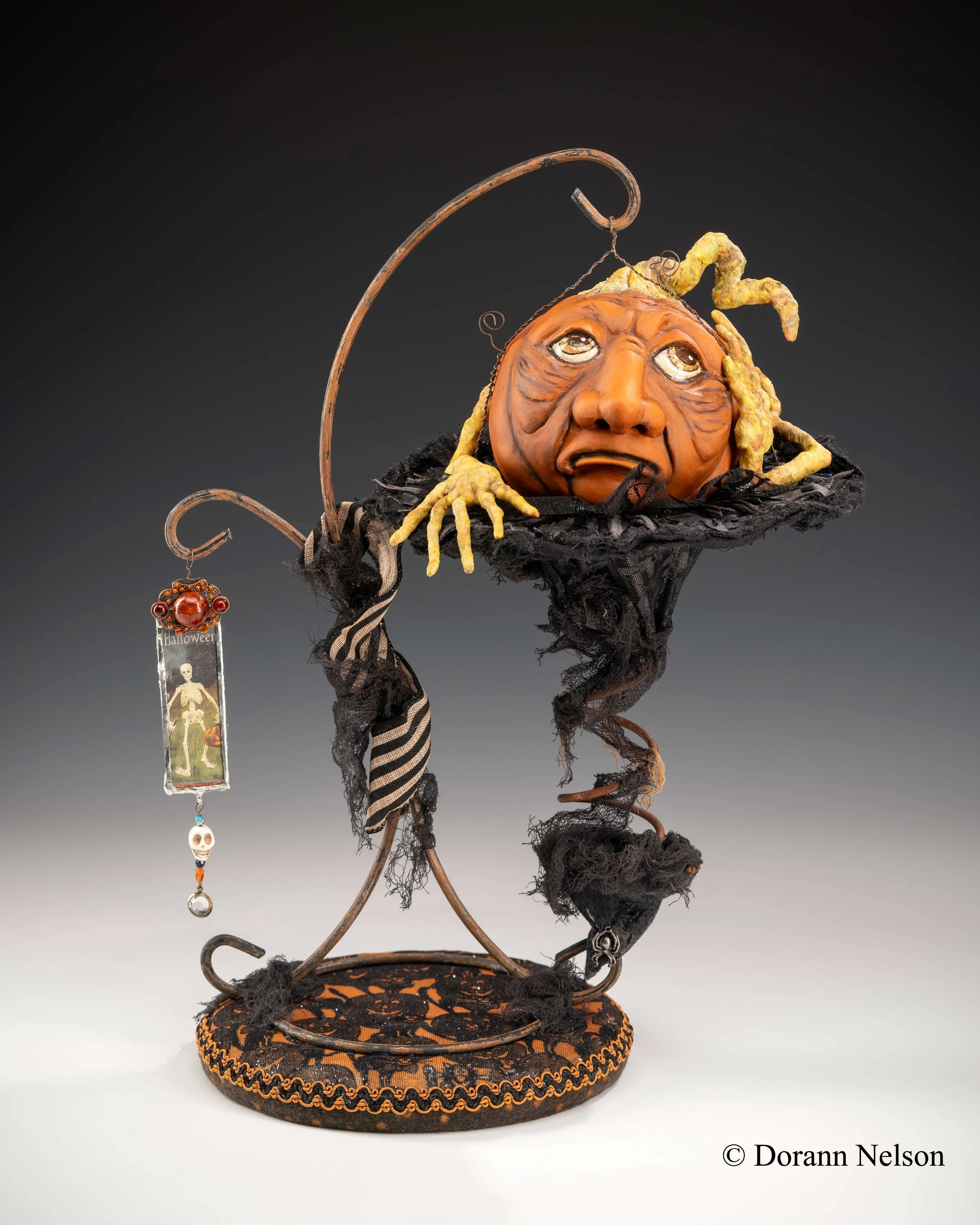 An art piece depicting a witch with a pumpkin head, featuring a sad expression, resting on a black decorated base, with a hook holding a Halloween-themed charm. The art uses black fabric, striped ribbon, and twisted metal to create a whimsical Hallow