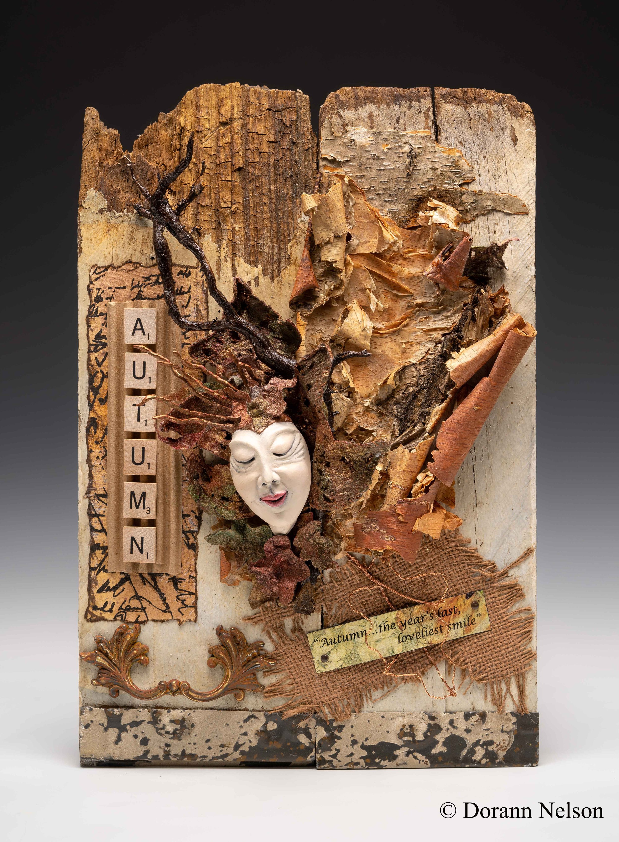 Mixed media artwork featuring a face mask, autumn leaves, wood pieces, and decorative elements, with a label reading 'Autumn...the year's last, loveliest smile'.
