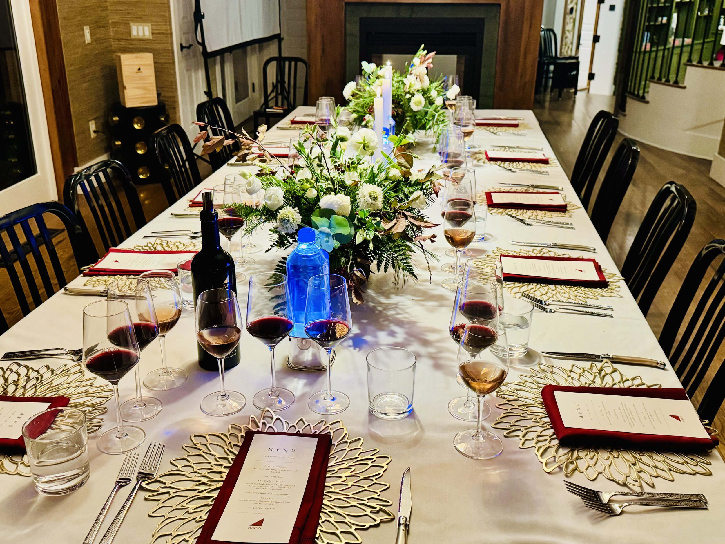 A long rectangular dining table set for a formal dinner with floral centerpiece, wine glasses, bottles of wine, menus, and gold decorative placemats in a well-lit room.
