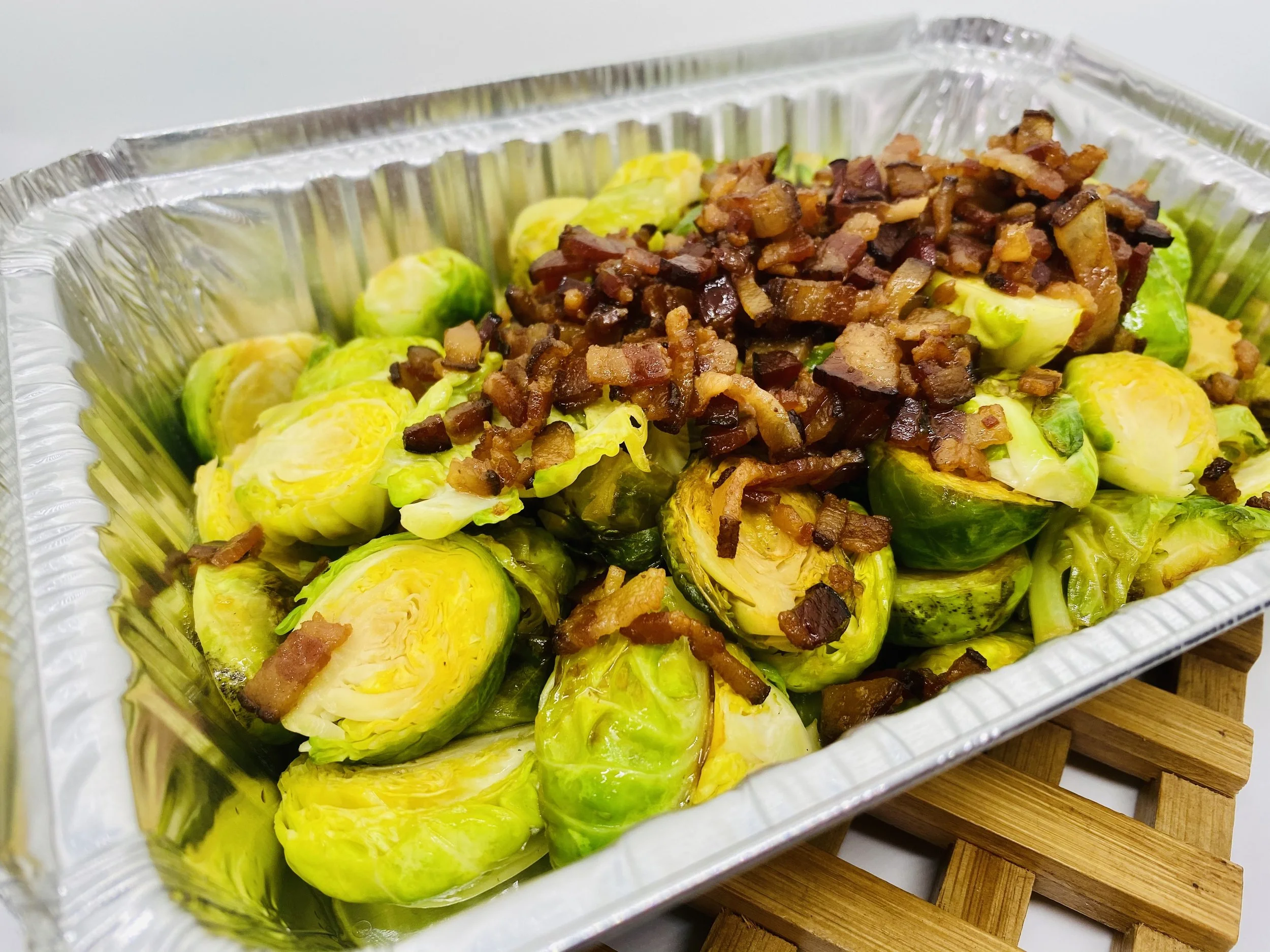 Cooked Brussels sprouts topped with crispy bacon in a foil tray.