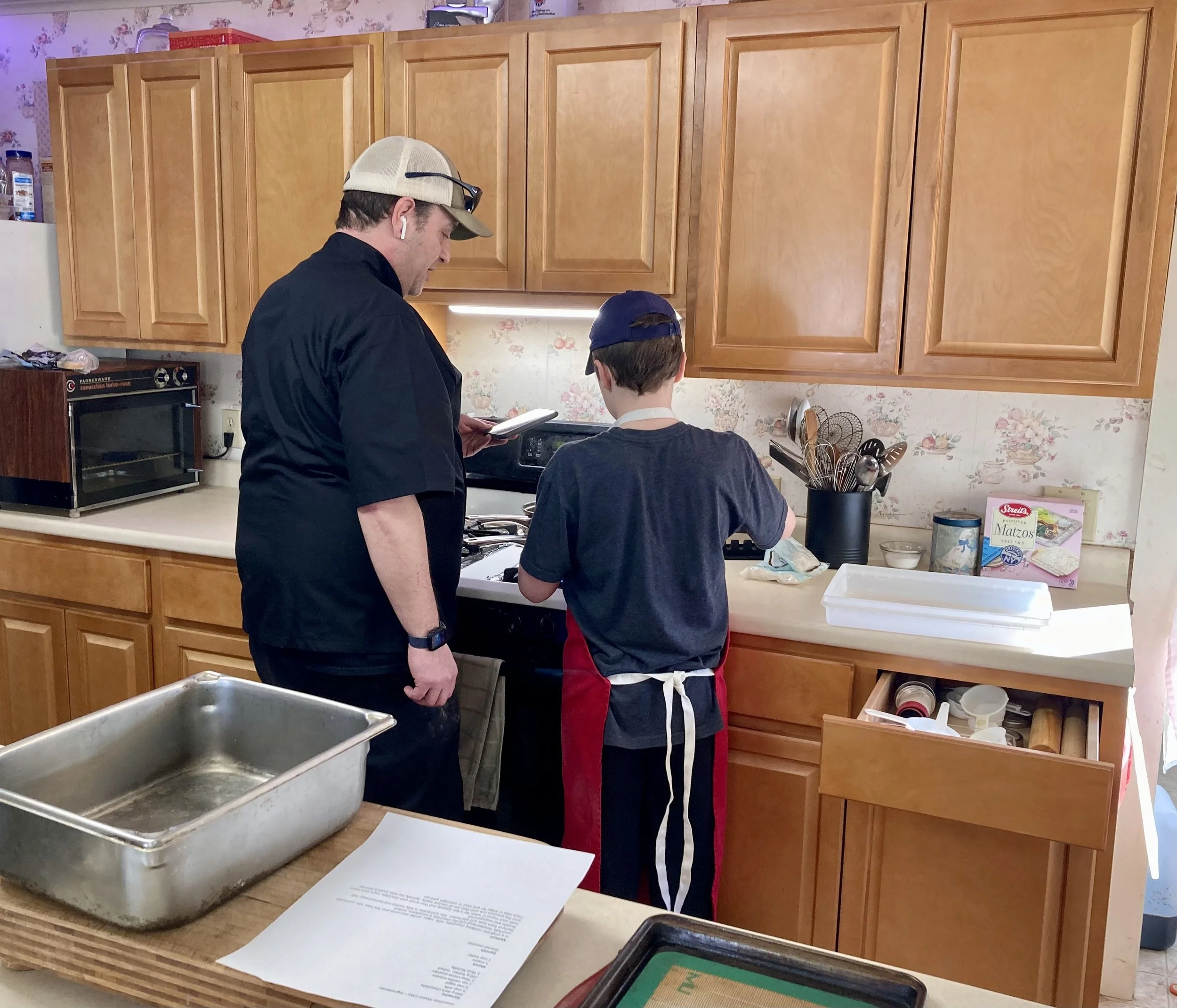 A man and a boy cooking together in a kitchen. The man wears a beige cap and AirPods, holding a notepad, while the boy, wearing a blue cap and apron, is at the stove. The kitchen has wooden cabinets and various cooking items on the counters.