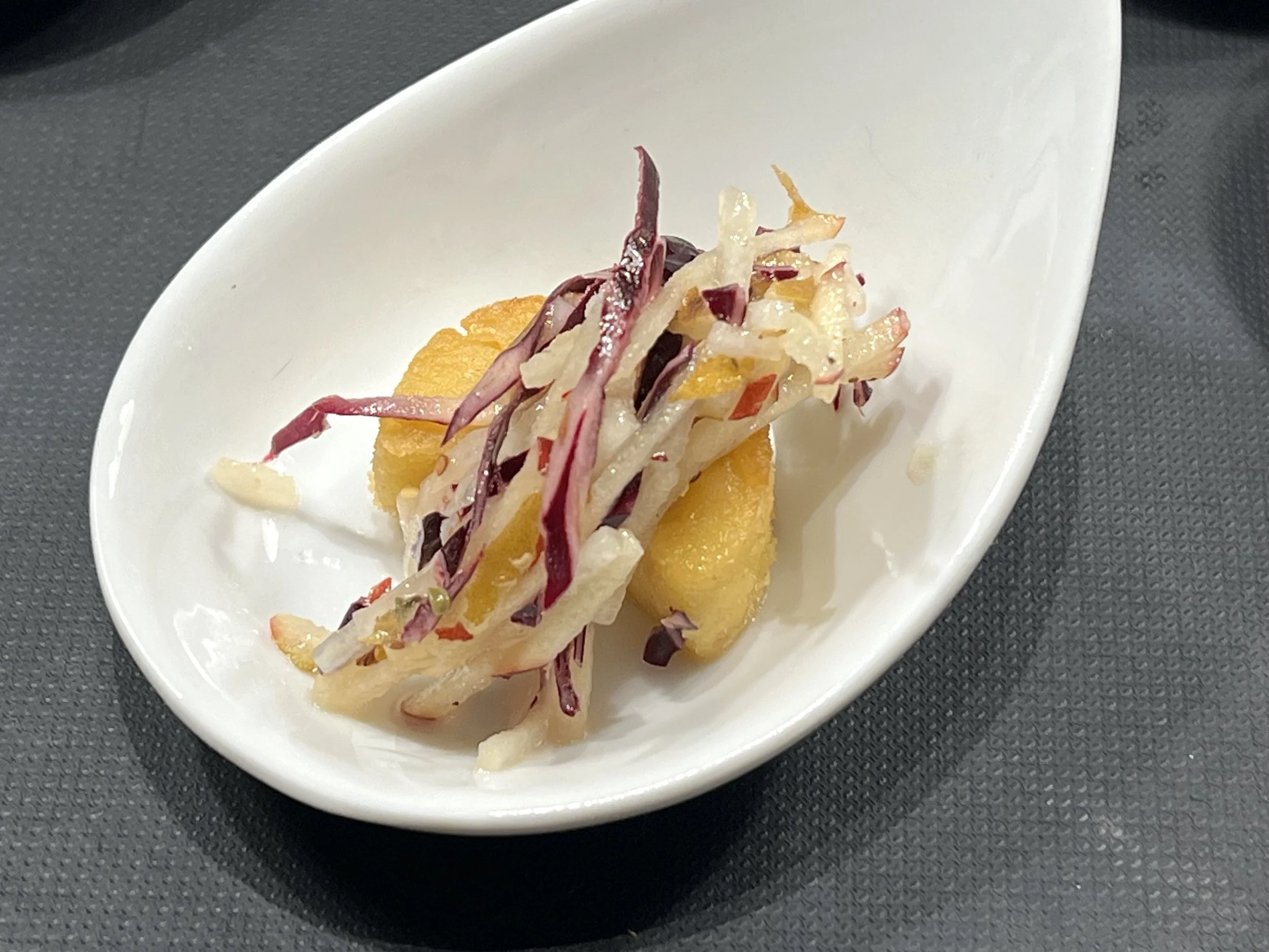 A white spoon holding a serving of creamy salad with shredded vegetables and a breaded item underneath, on a dark textured surface.