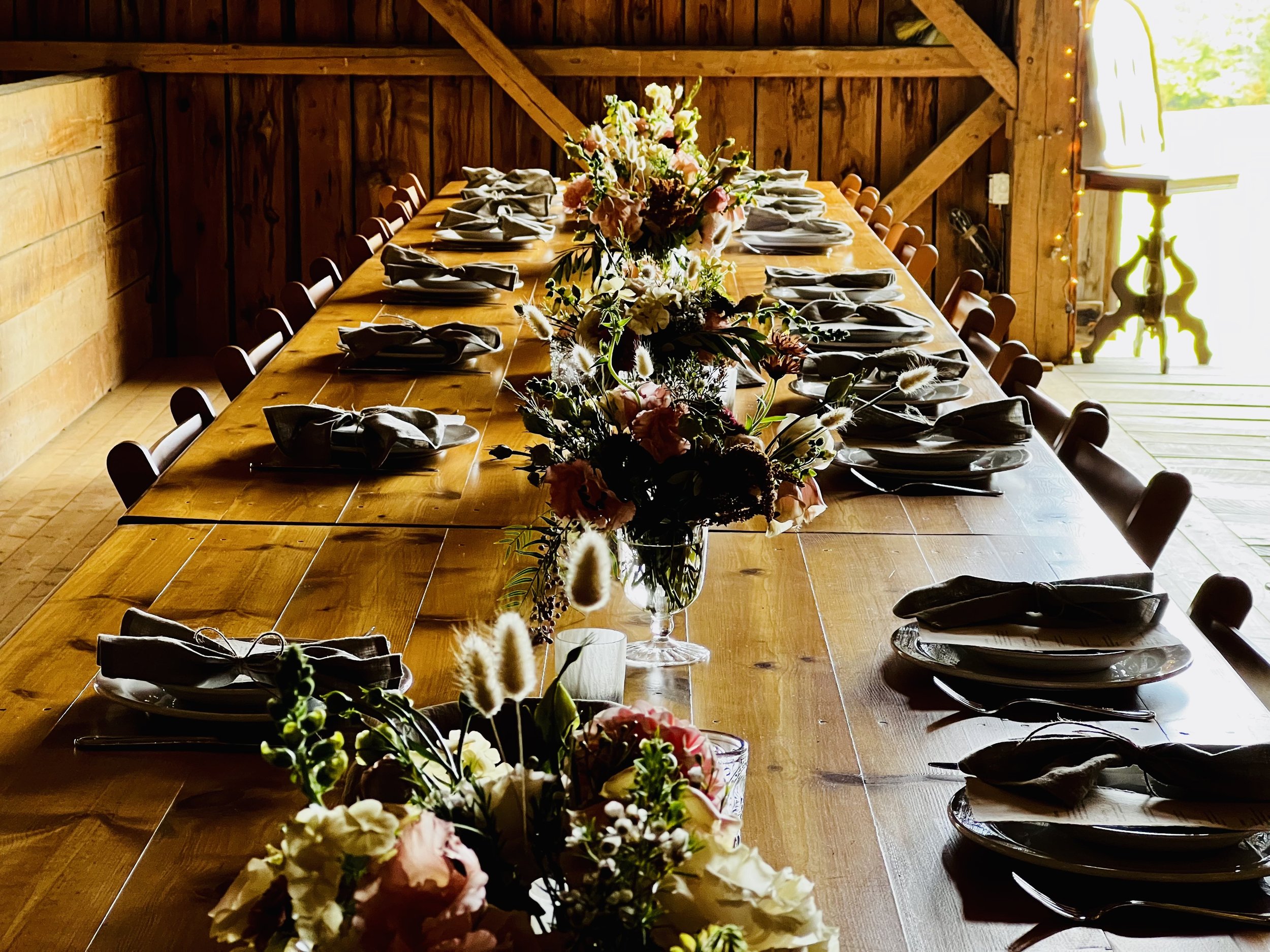 Long wooden table set for a meal with plates, silverware, and folded napkins, decorated with floral centerpieces in a rustic wooden room with sunlight coming through an open door.