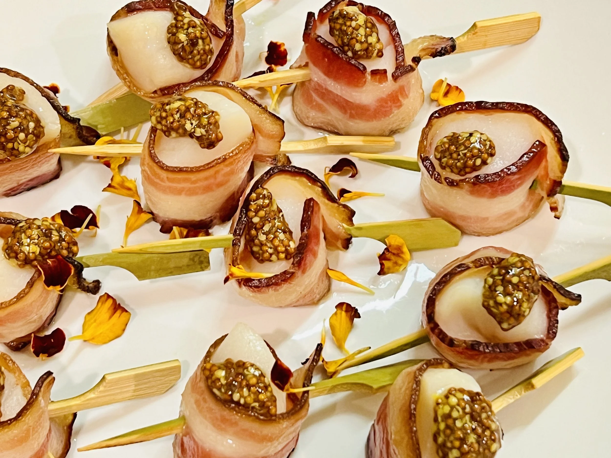 Bacon-wrapped pear bites topped with mustard seeds, skewered with bamboo sticks, garnished with flower petals.