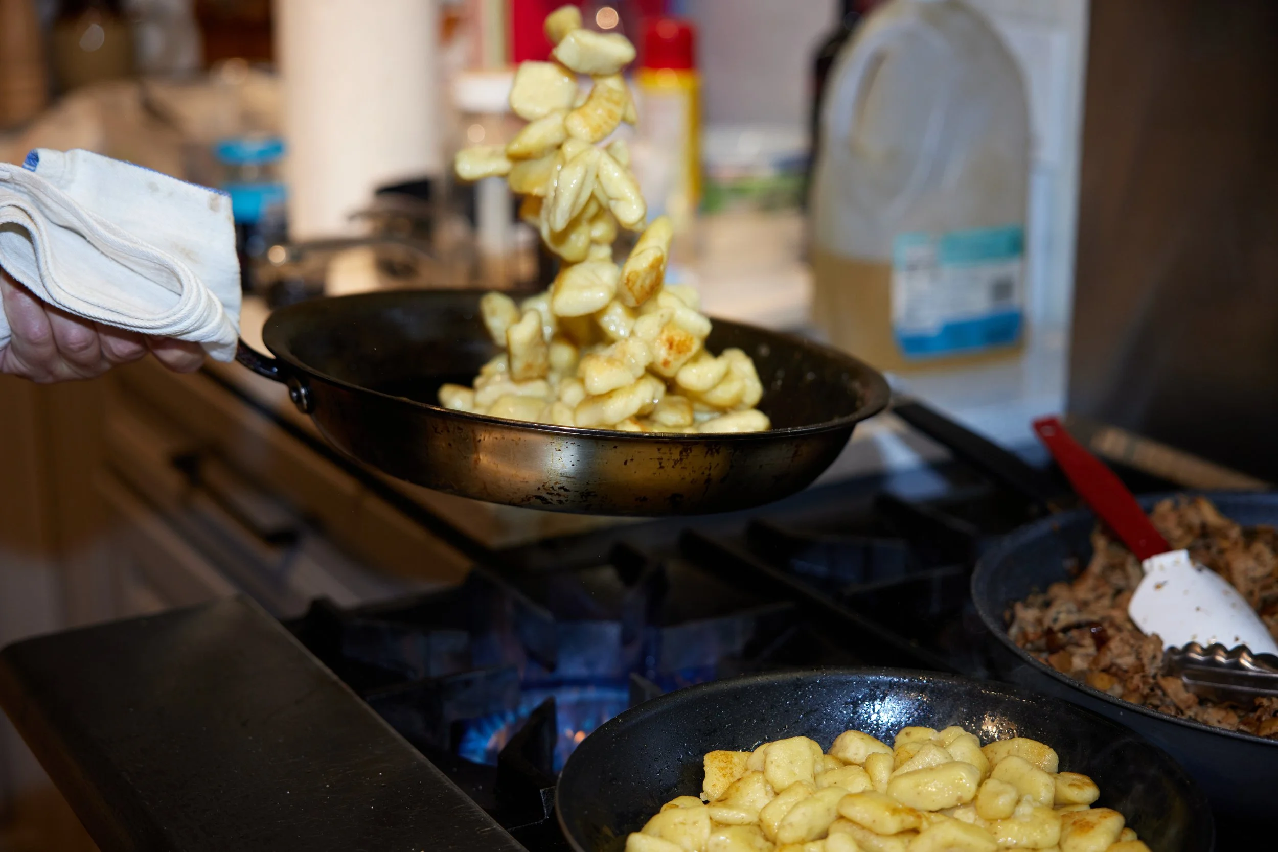 Person cooking on a stove, tossing gnocchi from a frying pan into a bowl, with another skillet containing cooked food nearby.