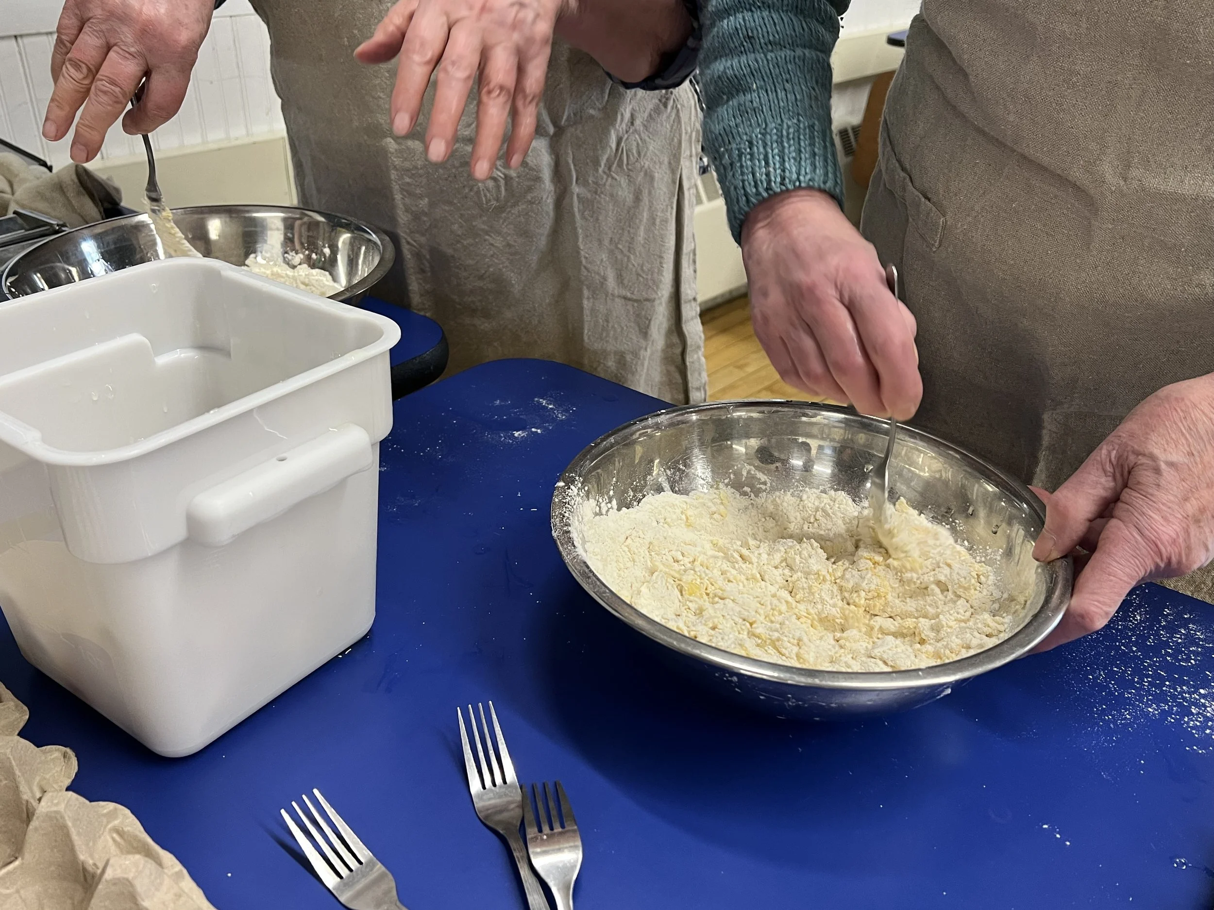 Two people mix ingredients in a stainless steel bowl, with kitchen utensils and a white container on a blue table.