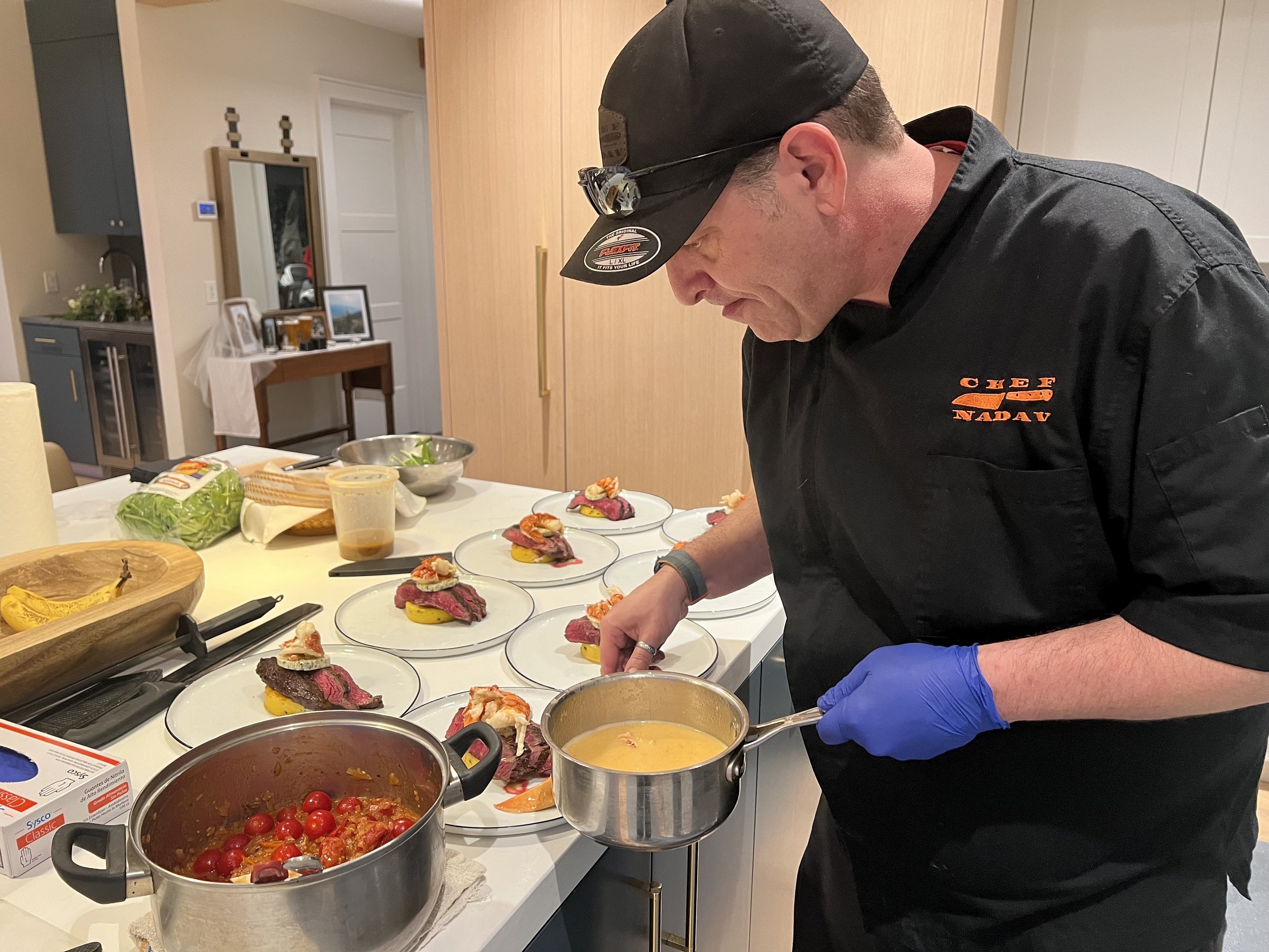 Chef in black uniform and cap preparing dishes with sliced beef, vegetables, and sauce on a kitchen counter, with plated appetizers in the background.