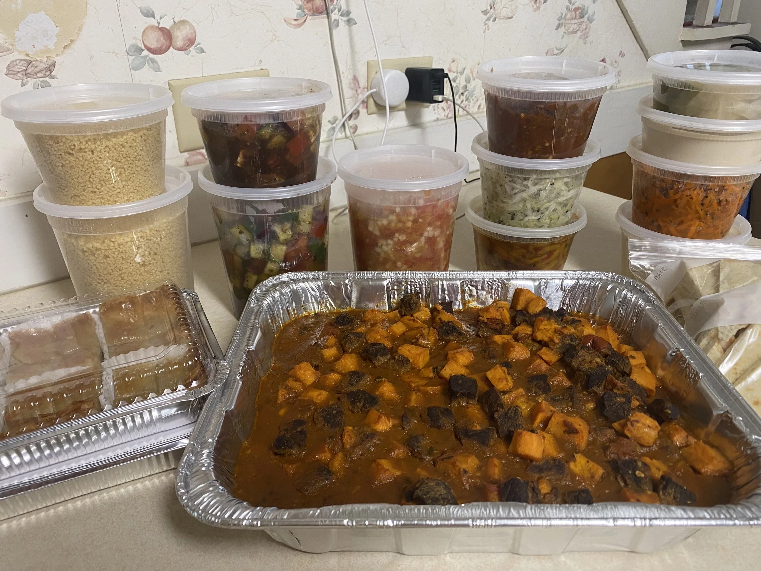 A large tray of sweet potato and black bean chili, several containers of chopped salads and side dishes, and a plastic container of tamales on a kitchen counter.