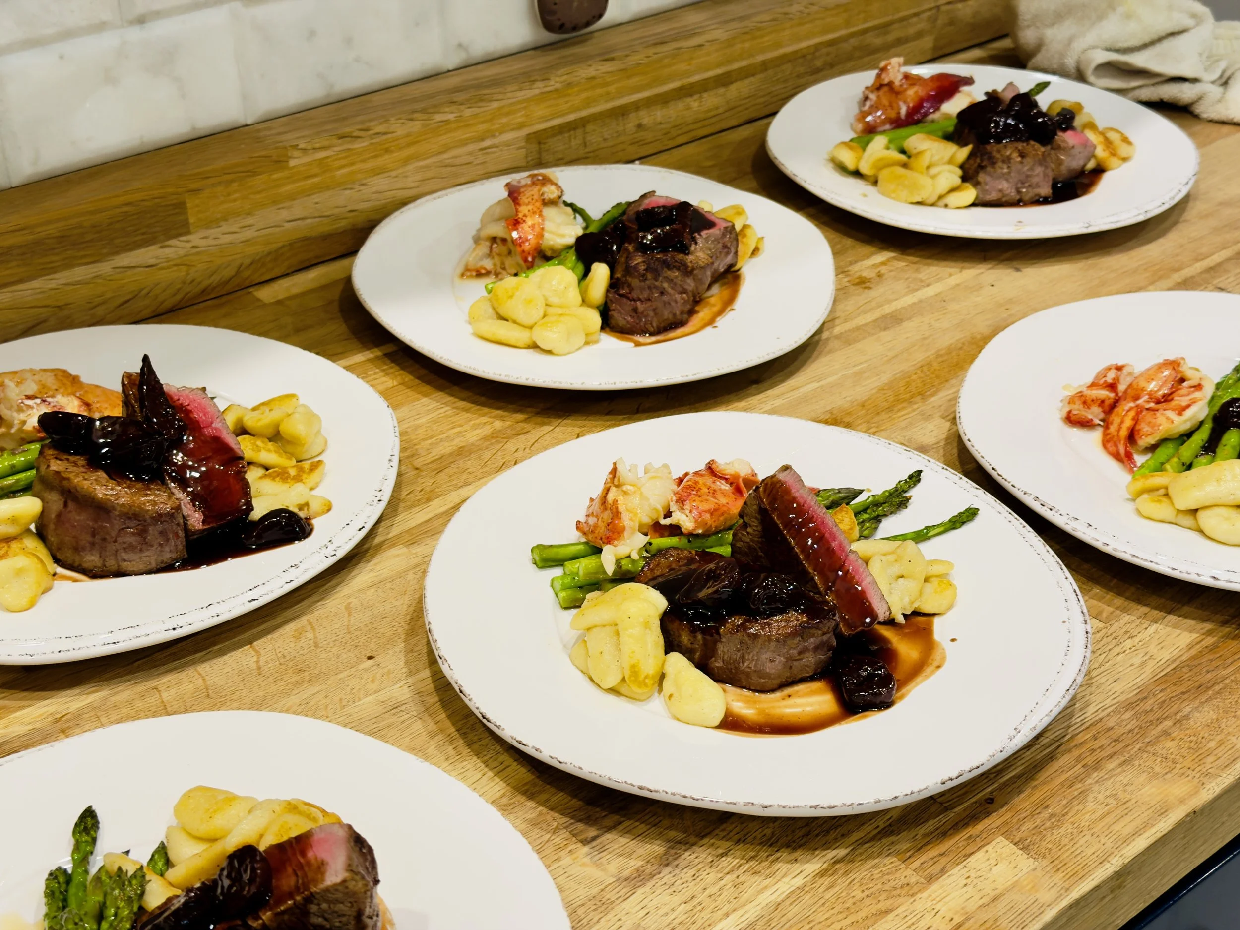 Five plates of cooked beef filet mignon with sauce, garnished with vegetables including asparagus, gnocchi, and a side of lobster tail with sauce, on a wooden table.