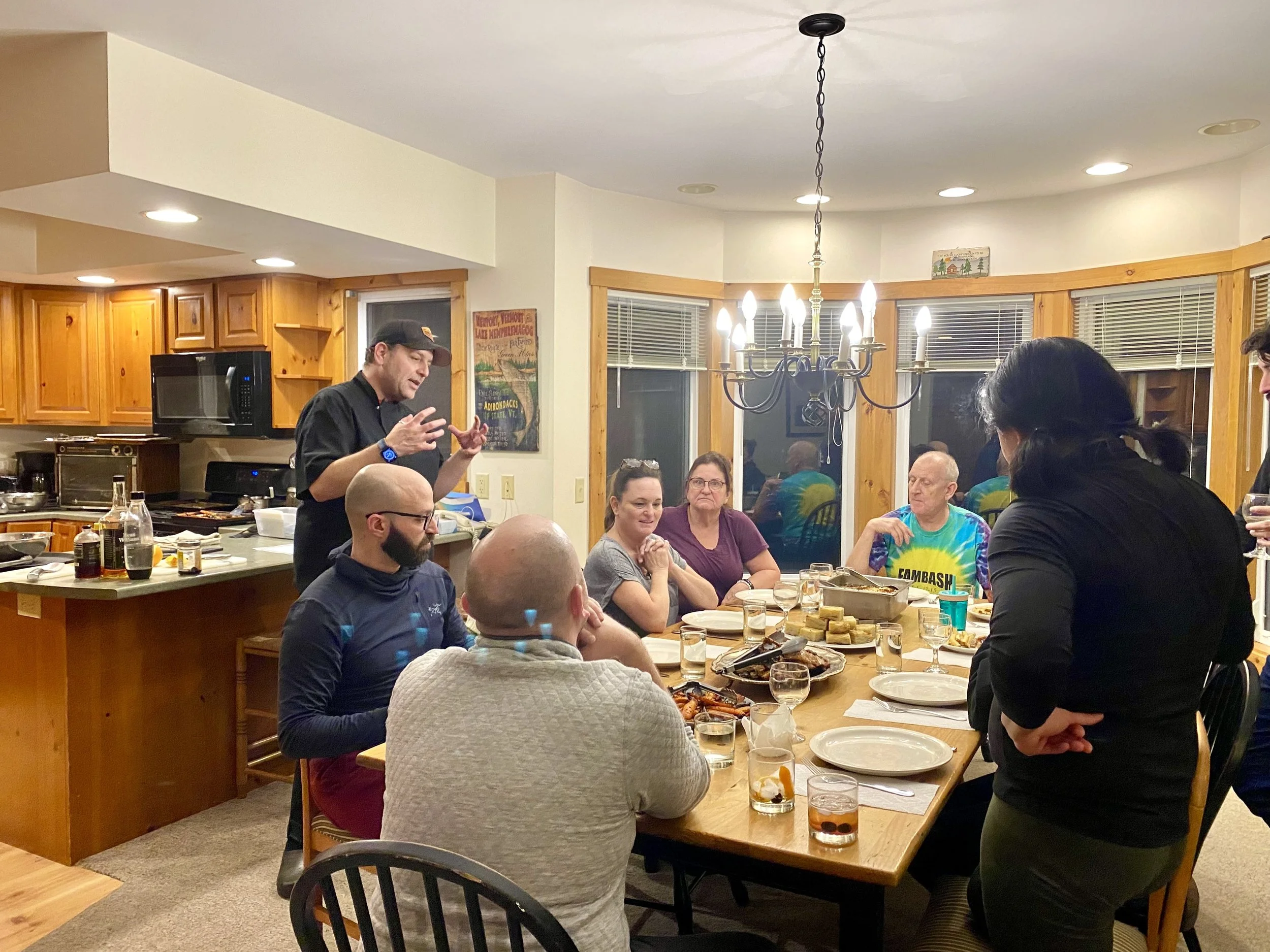 Group of people gathered around a dining table during a dinner party, with a chef speaking nearby, in a cozy kitchen and dining area with wooden accents and a chandelier.