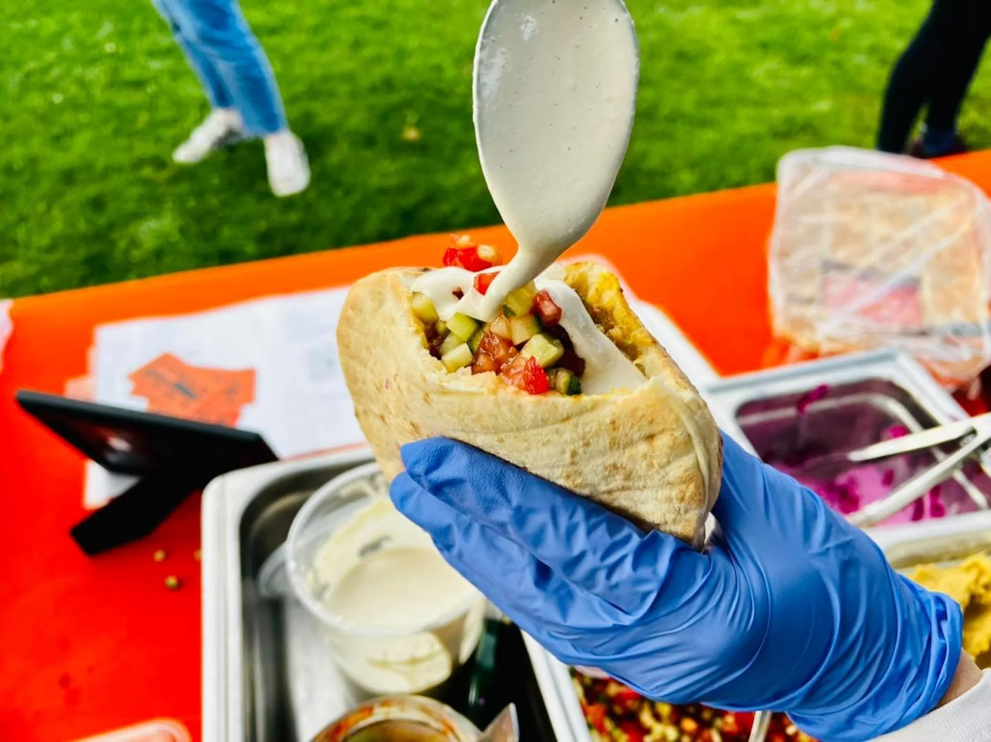 A person wearing a blue glove holding a loaded taco with toppings, pouring white sauce on it at an outdoor food stand.