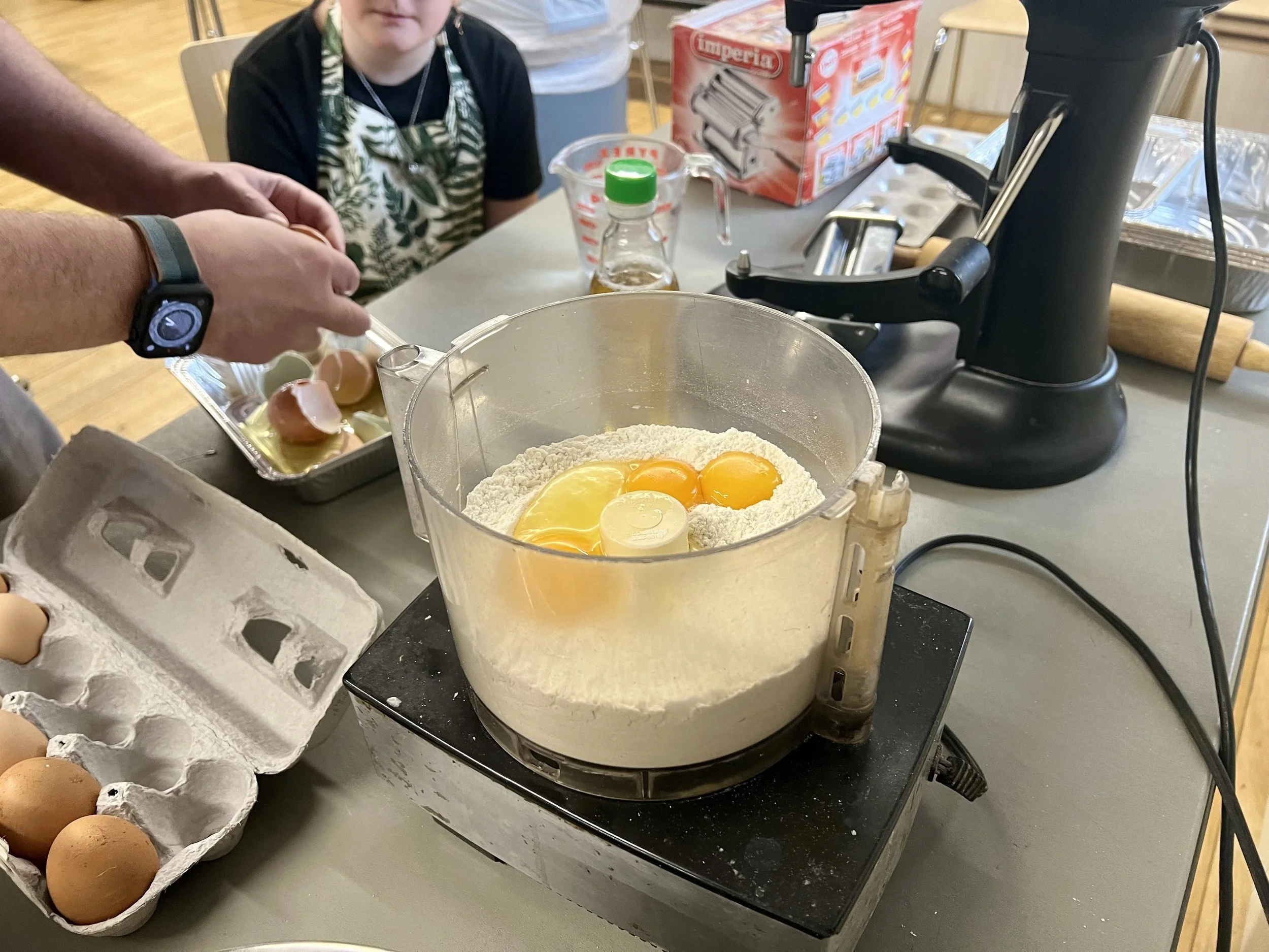 A person cracking eggs into a food processor with flour inside, on a kitchen countertop with eggs, a carton, and baking tools nearby.