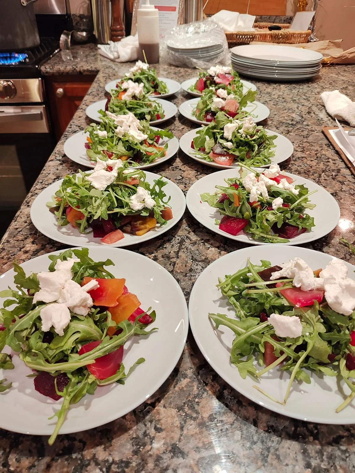 Multiple plates of mixed green salad with cherry tomatoes, beetroot, and white cheese on a granite kitchen counter.