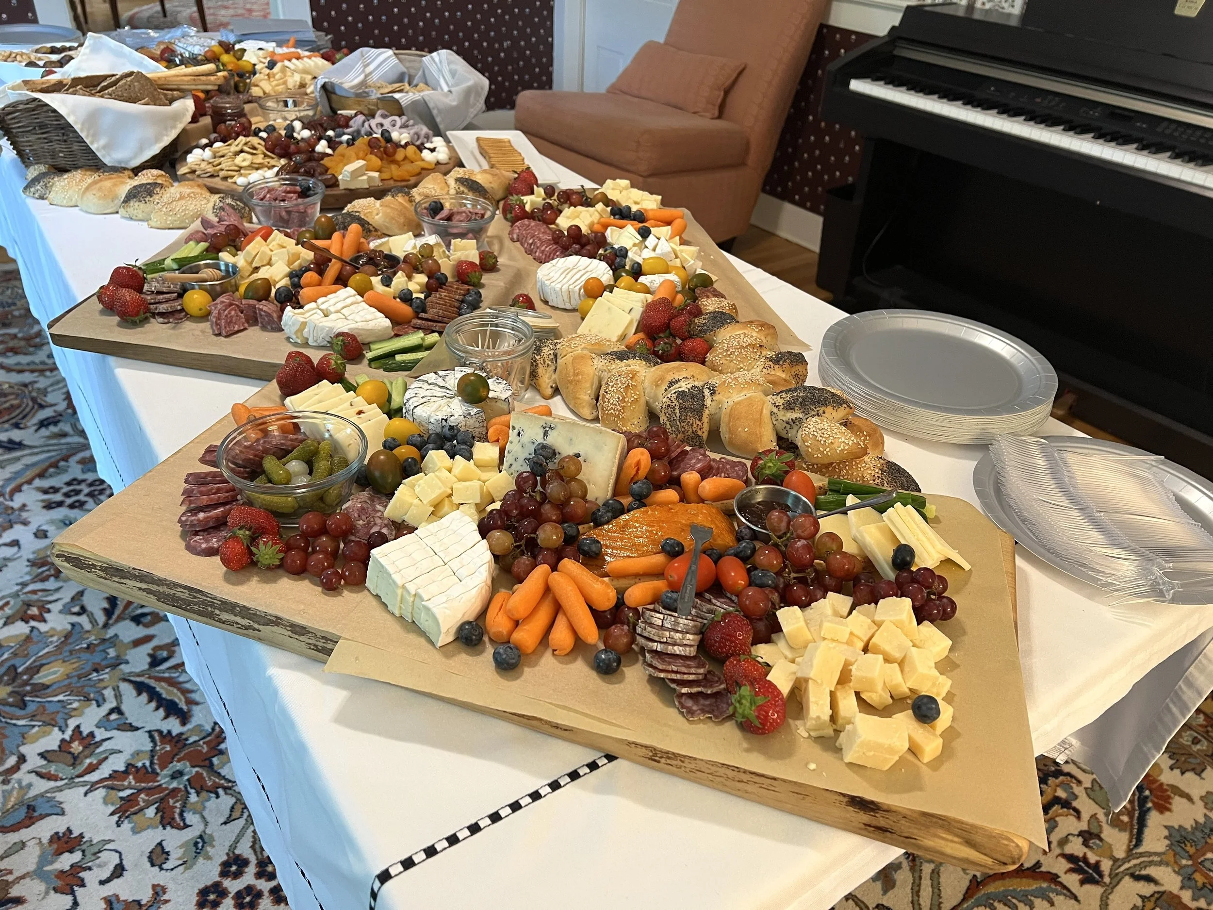 Table with assorted cheeses, meats, fresh fruits, vegetables, bread rolls, and snacks for a party or gathering. Plates and napkins are also visible.