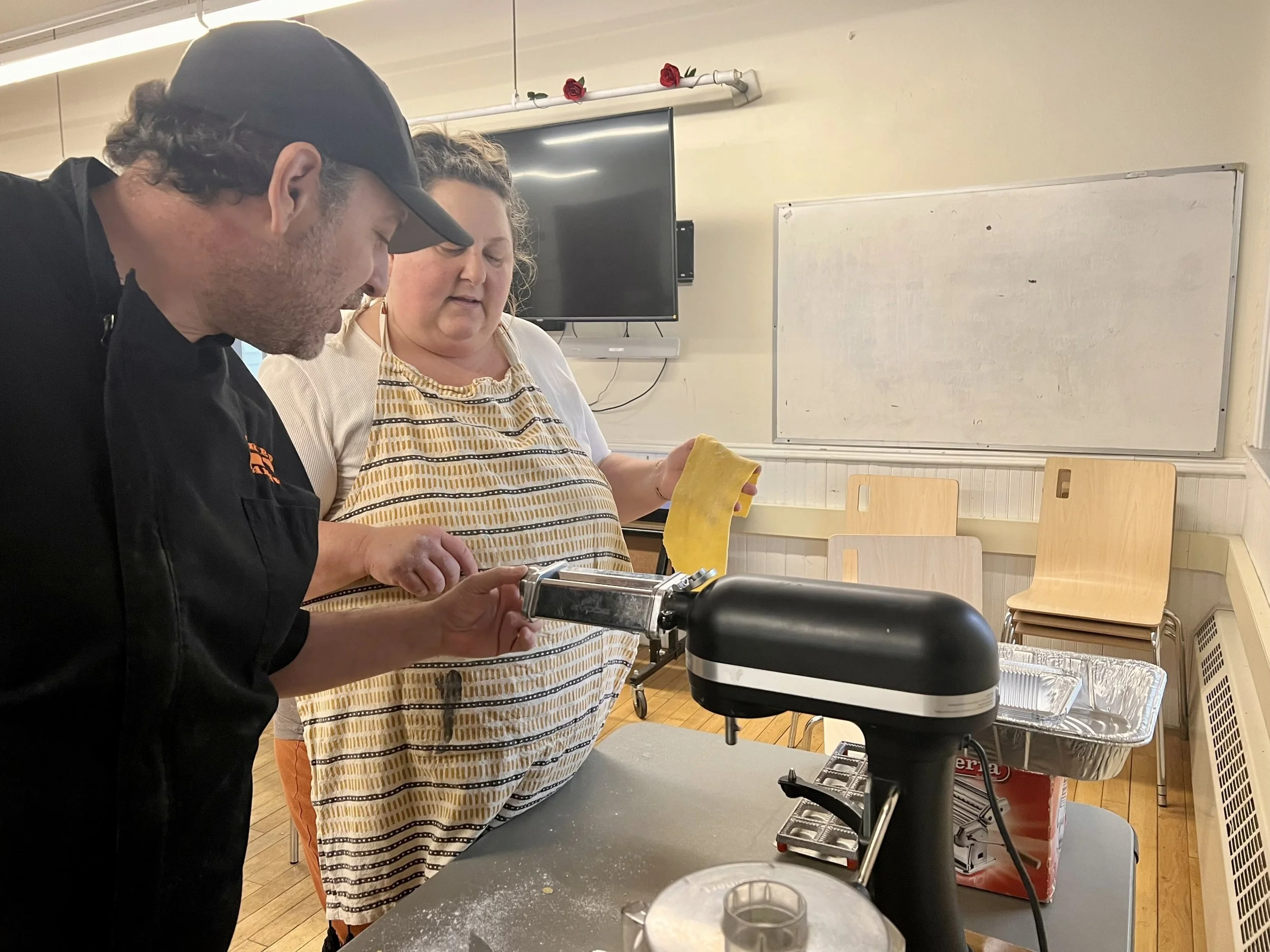 Two people working together in a kitchen, one holding a pasta machine and the other holding a piece of dough. There are chairs stacked in the background and a whiteboard on the wall.