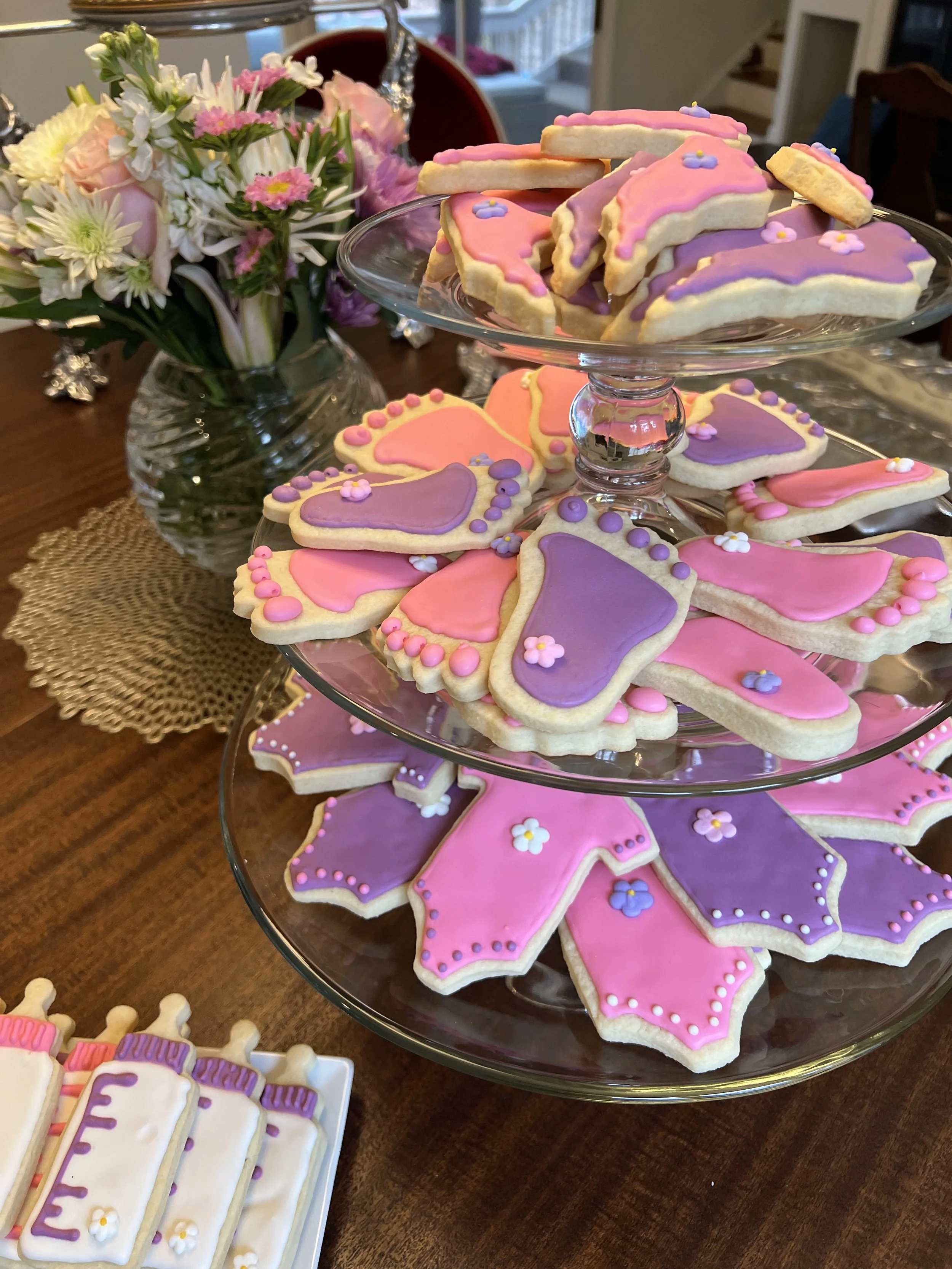 Tiered glass cake stand with pink and purple decorated cookies on a wooden table, with a floral arrangement in a glass vase in the background.