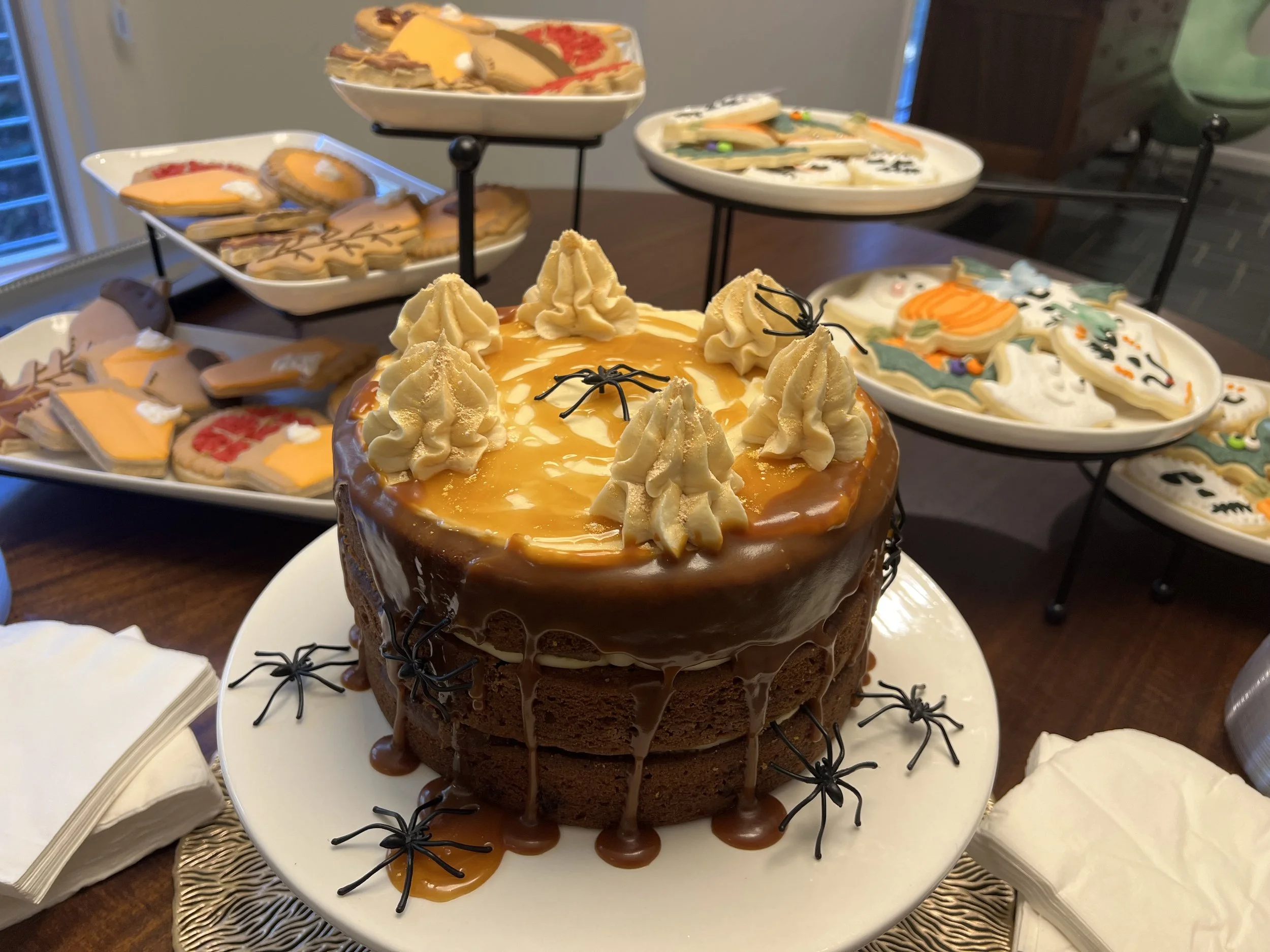 Chocolate cake decorated with caramel sauce, whipped cream rosettes, and black spider decorations, surrounded by Halloween-themed cookies and treats.