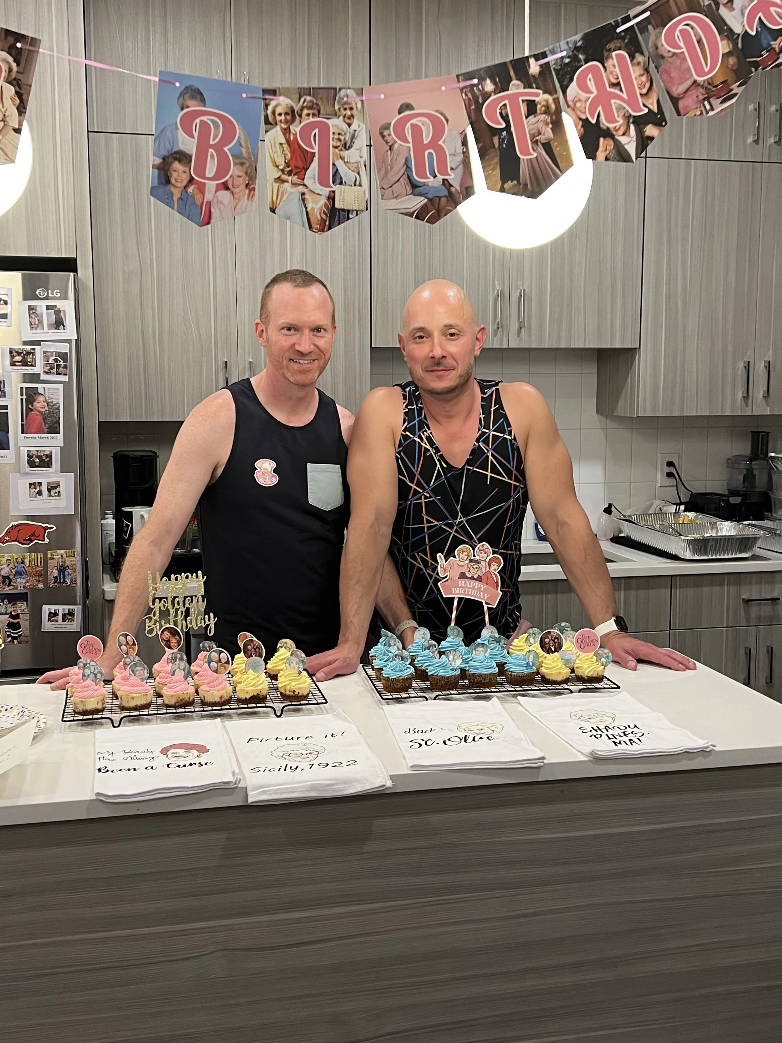 Two men celebrating a birthday with decorated cupcakes on a kitchen counter, birthday banners hanging above, and birthday-themed napkins.