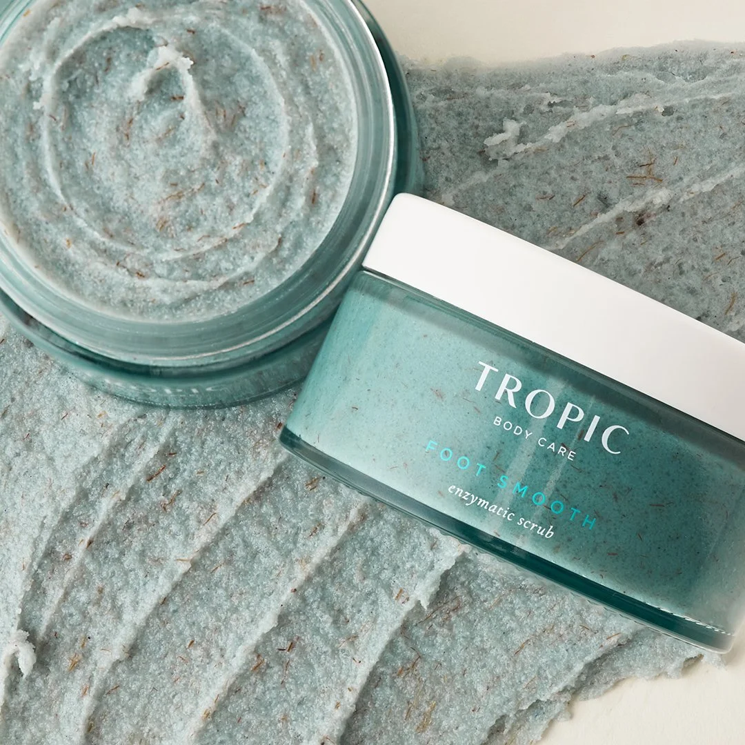 A jar of tropical foot smoothing enzymatic scrub with a teal label, placed next to a pile of the scrub on a gray textured surface.