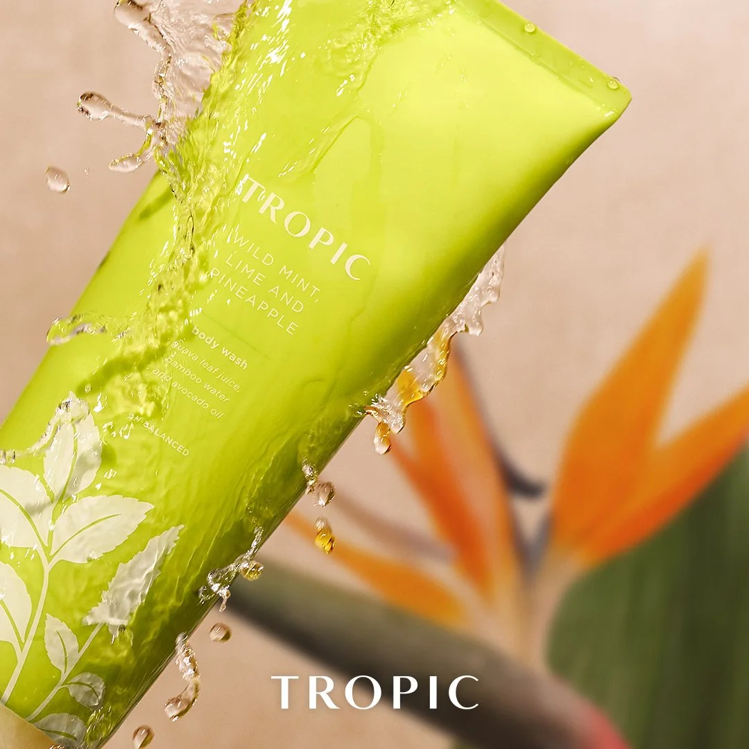 Green bottle of Tropic body wash labeled 'Wild Mint, Lime, and Pineapple' with water splashing around it, and a blurred background of tropical flowers.