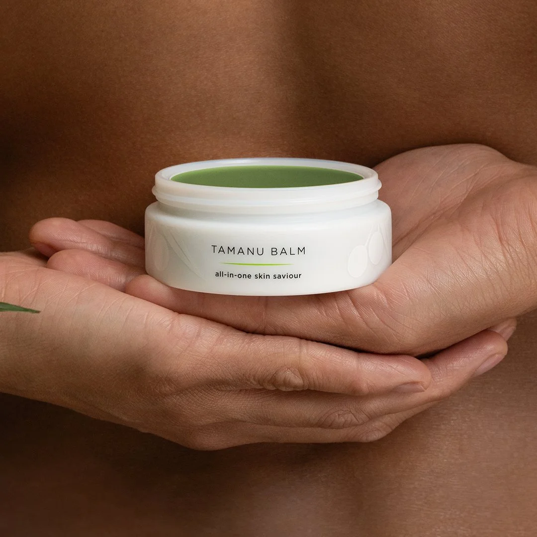 A person's hand holding an open jar of Tamanu Balm with a green balm inside, against a background of skin.
