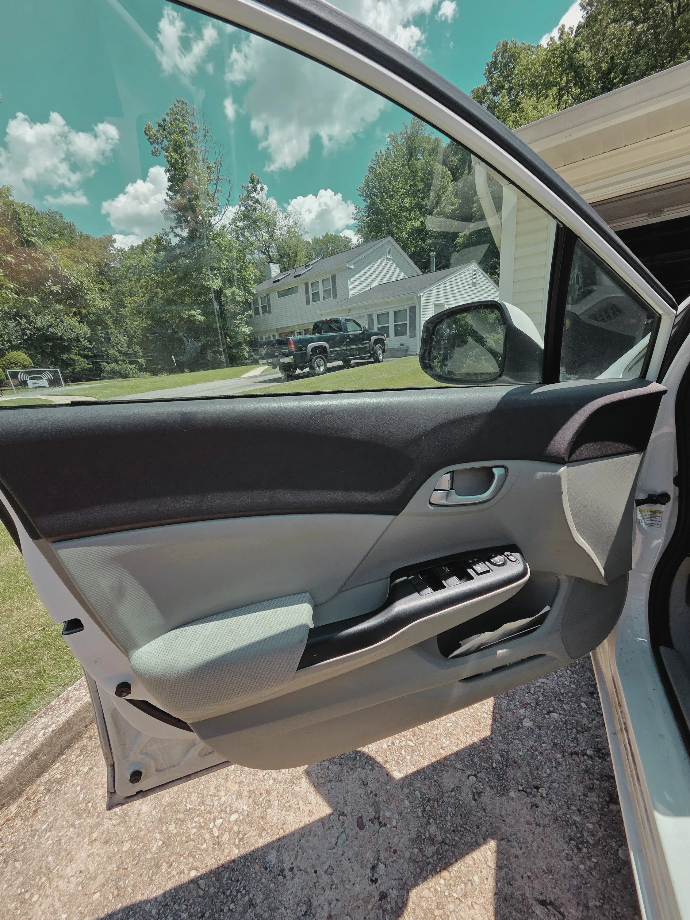 The interior of a white car with the front passenger door open, showing the gray and white interior, the window controls, and a view outside of a residential neighborhood with a black pickup truck, a white house, and trees under a partly cloudy sky.