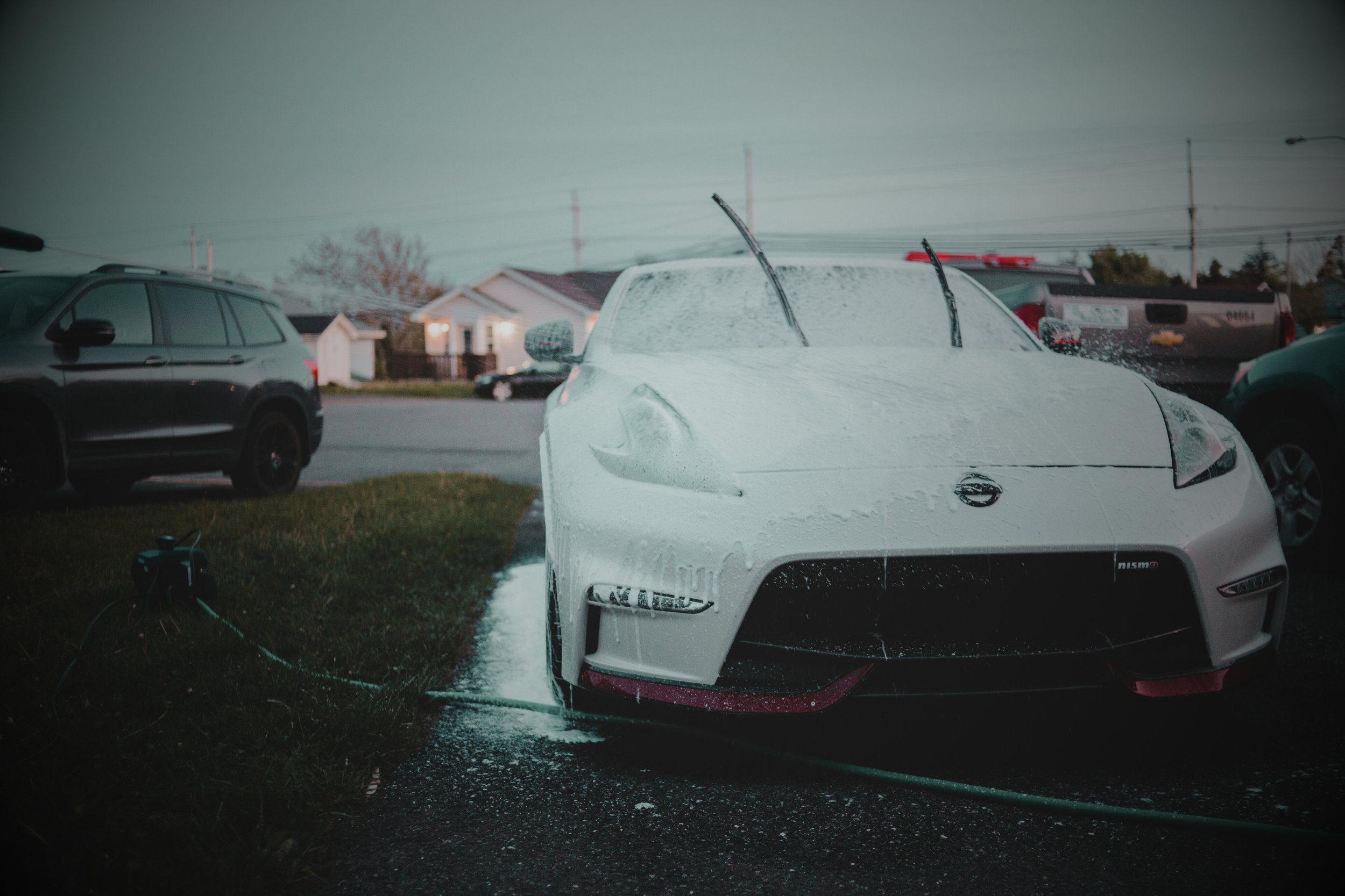 A white Nissan sports car being washed with soap and water, soap suds on the hood, windshield wipers raised, parked near a house and other vehicles.