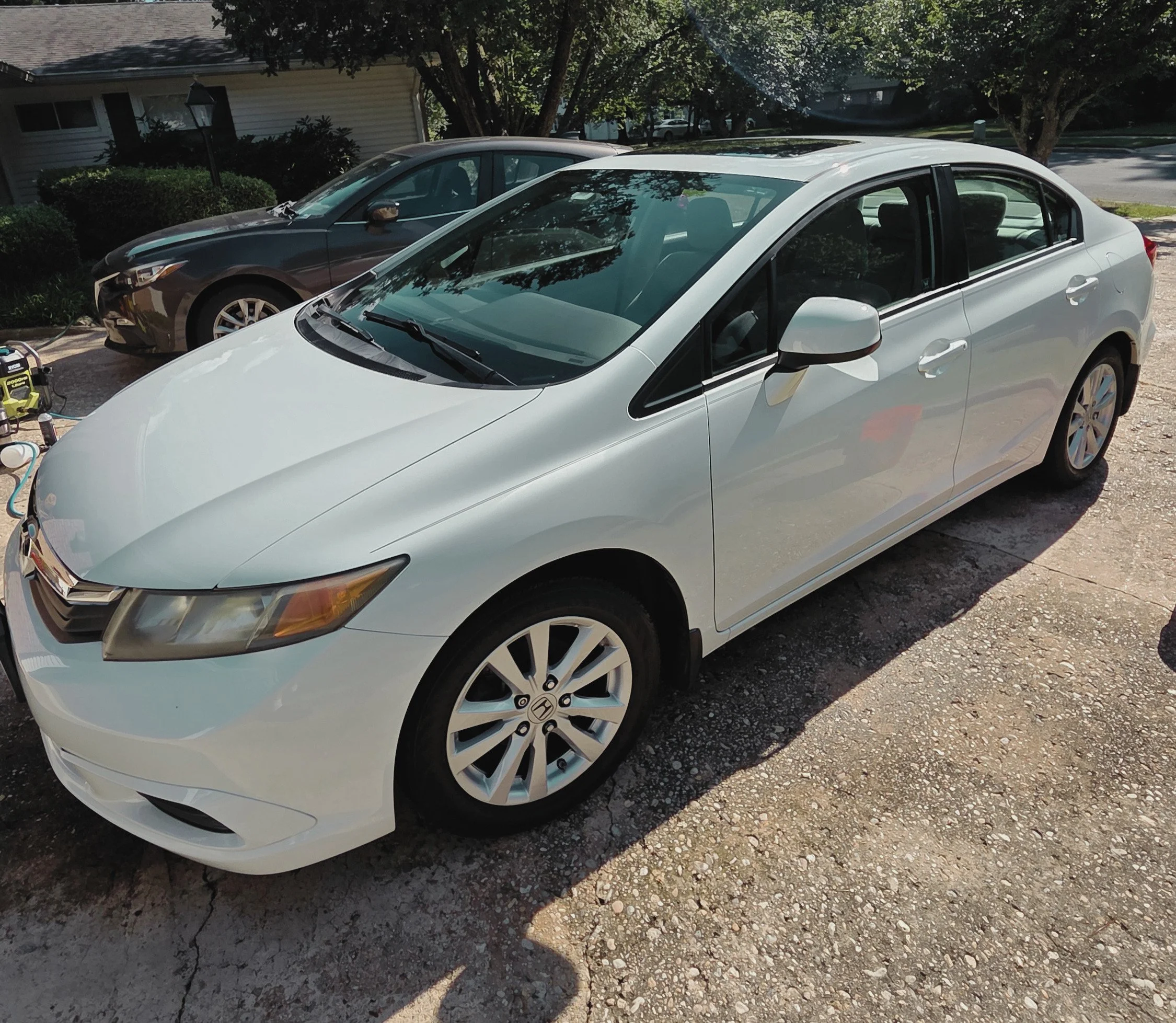 A white sedan and a gray car parked side by side on a gravel driveway in front of a house surrounded by trees and bushes.