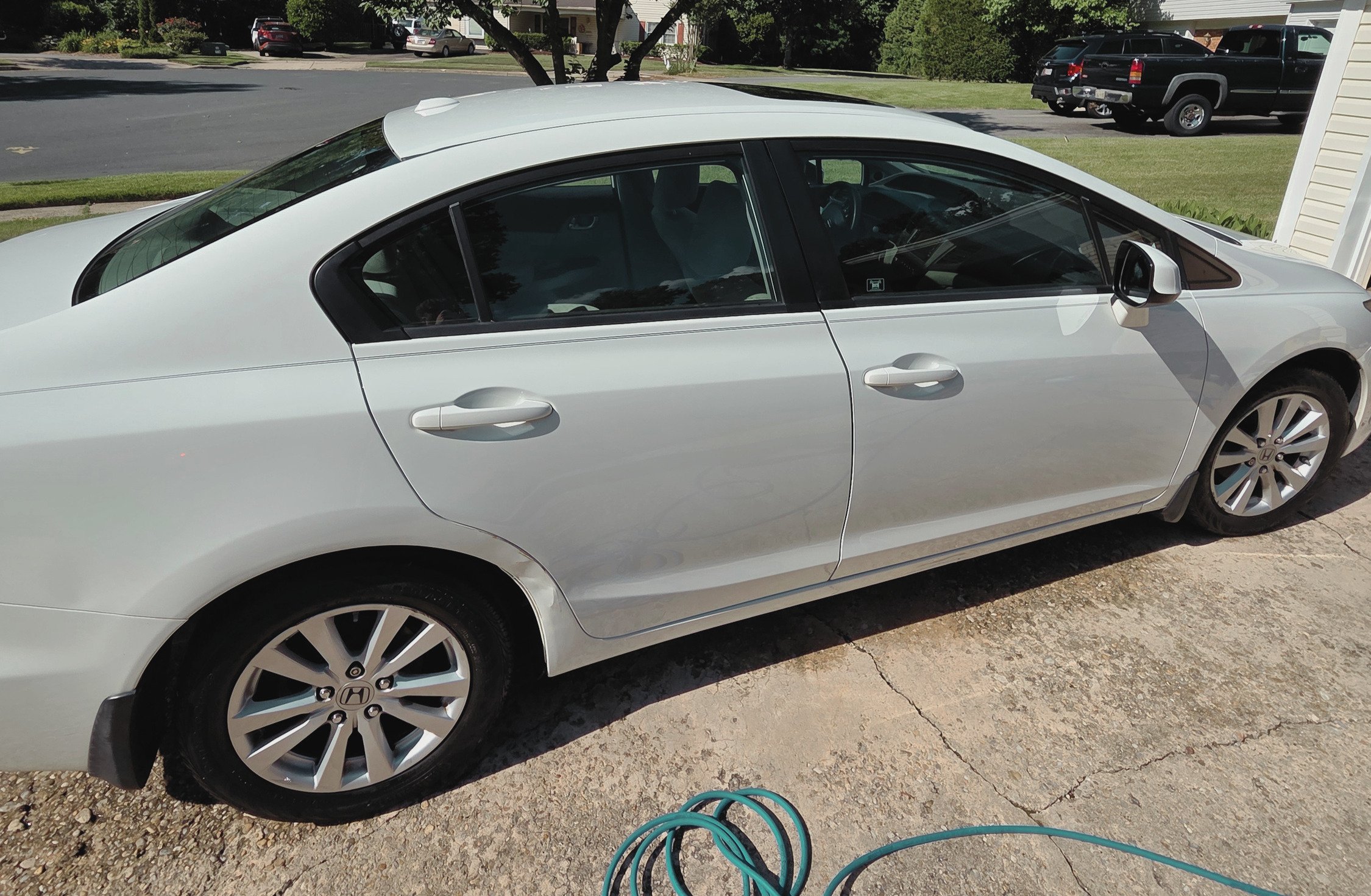 A white Honda sedan parked outdoors on a driveway with a green garden hose coiled on the ground in front of it. In the background, there are trees, other parked cars, and a house.
