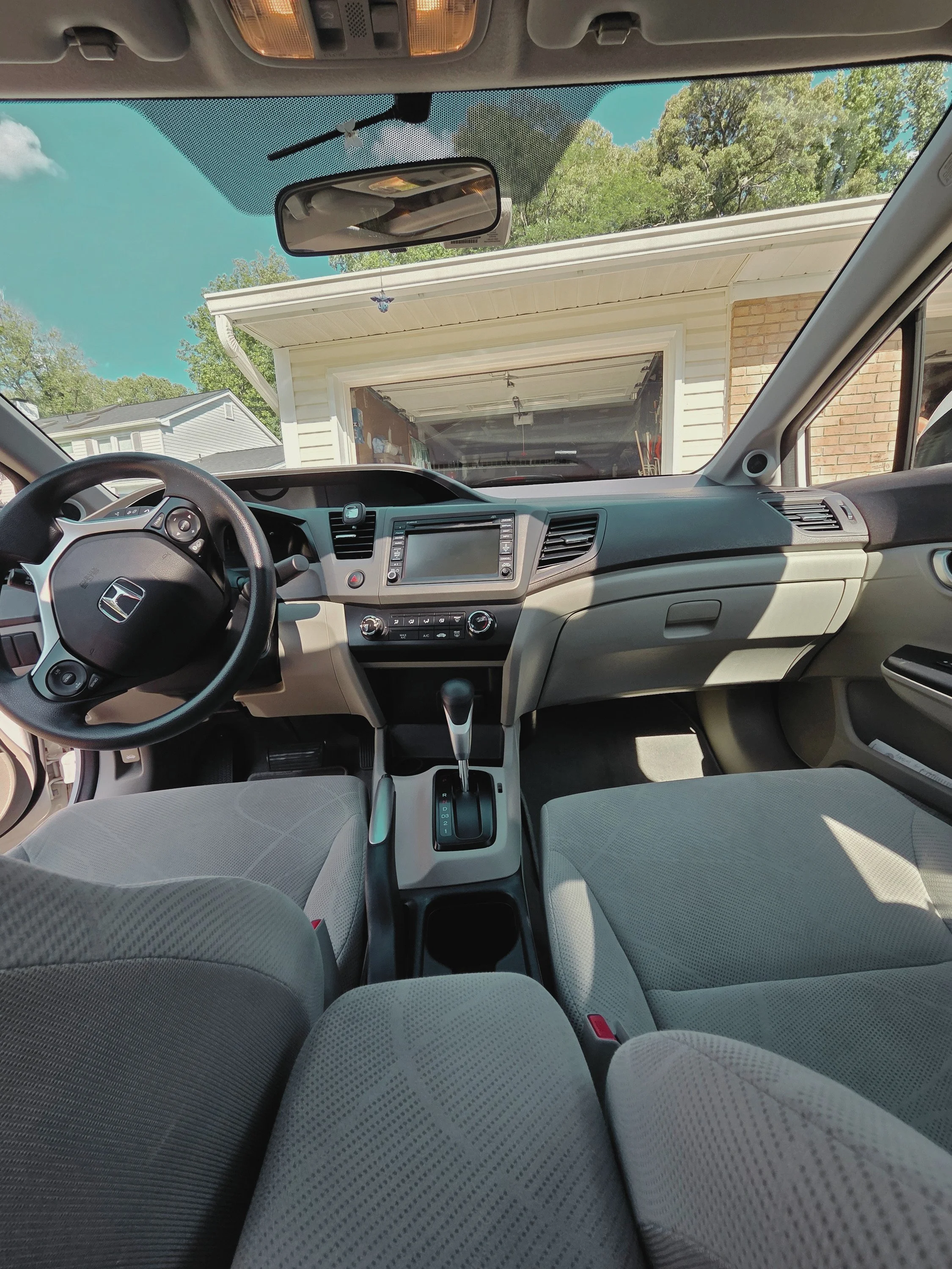 The interior of a car showing the front seats, dashboard, steering wheel, and gear shift. The view outside the windshield includes a garage and green trees under a blue sky.