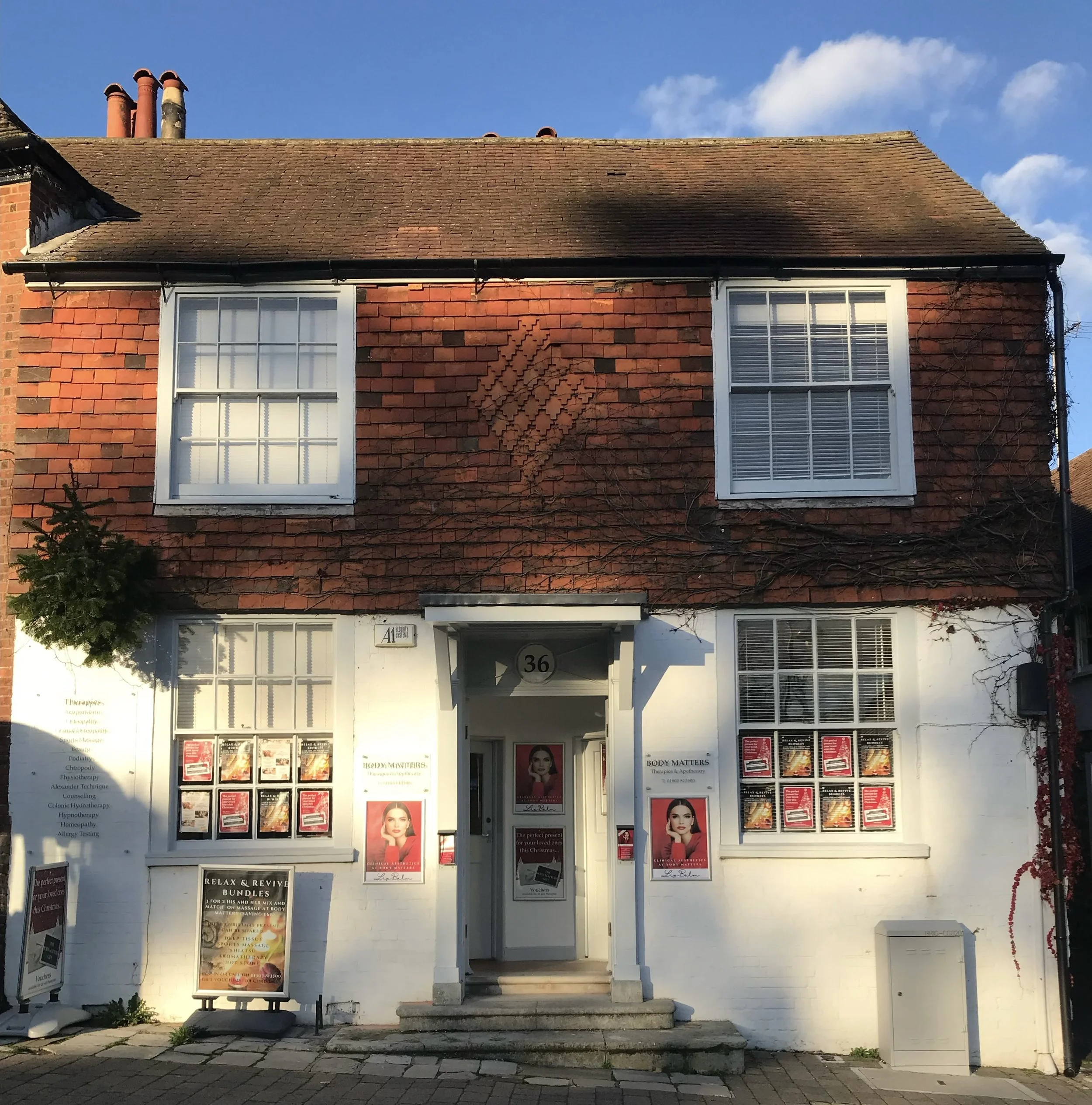 A two-story building with a brick upper facade and a white lower facade, featuring four large windows with white frames, a small front steps entrance, and signs advertising body therapy services outside.
