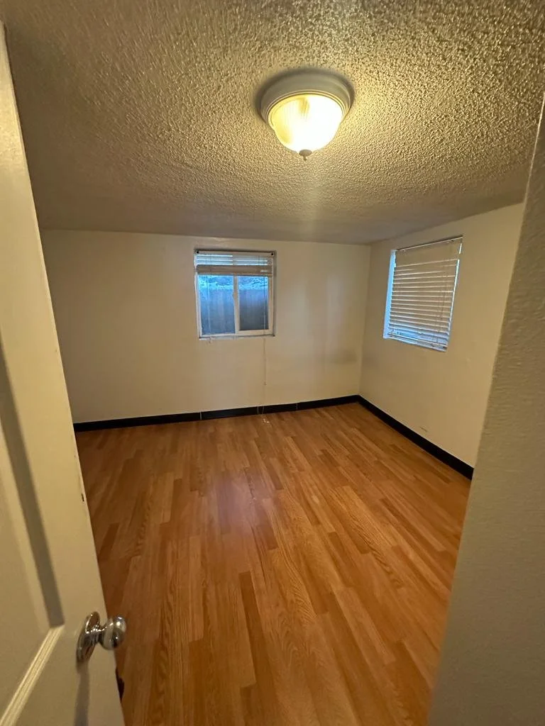 Empty small room with wooden floors, two windows with blinds, and an overhead light fixture.