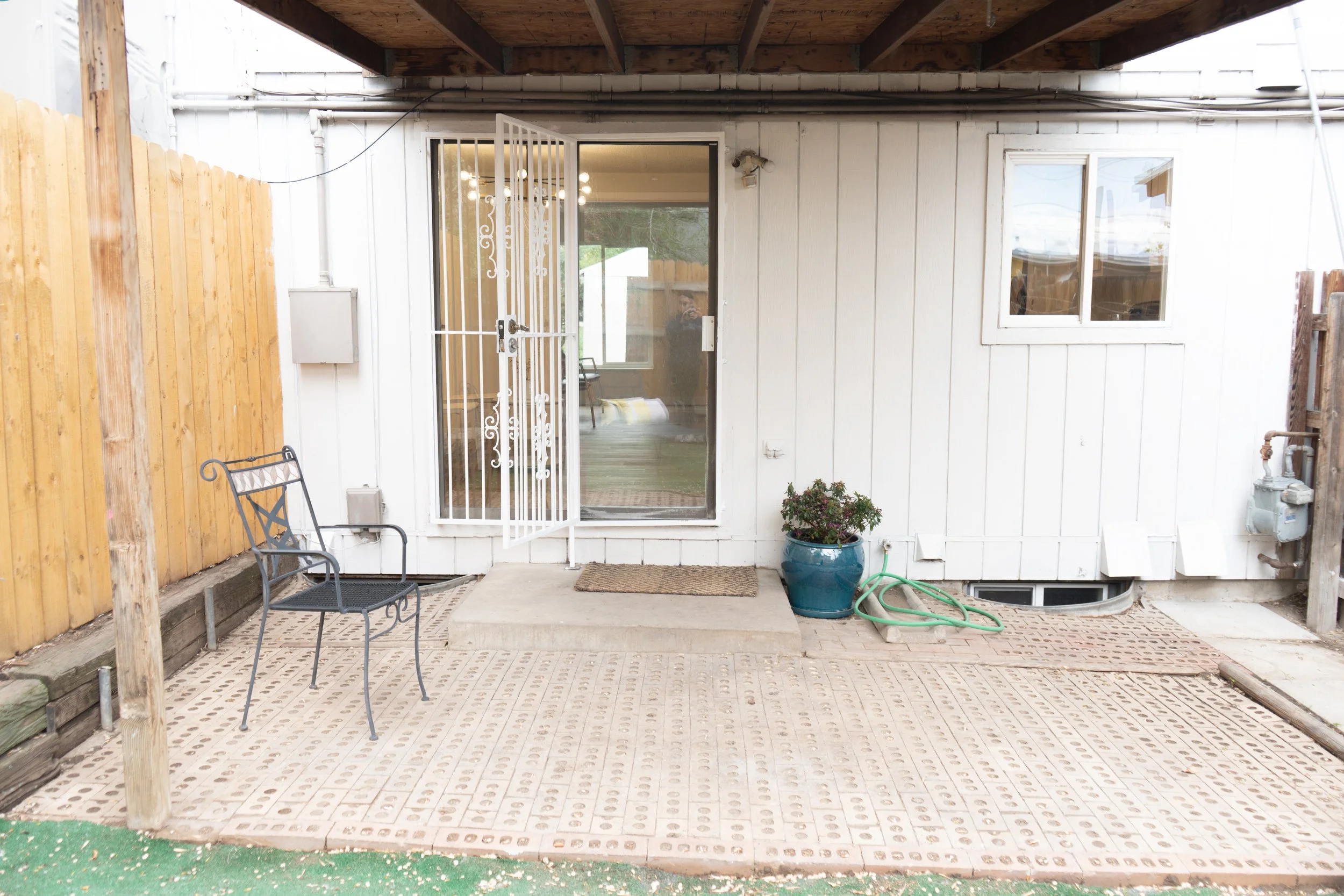 Back patio area with a black metal chair, a blue potted plant, a green garden hose, and a white door with a security screen, leading into a living room with visible chairs and windows, enclosed by a wooden fence.