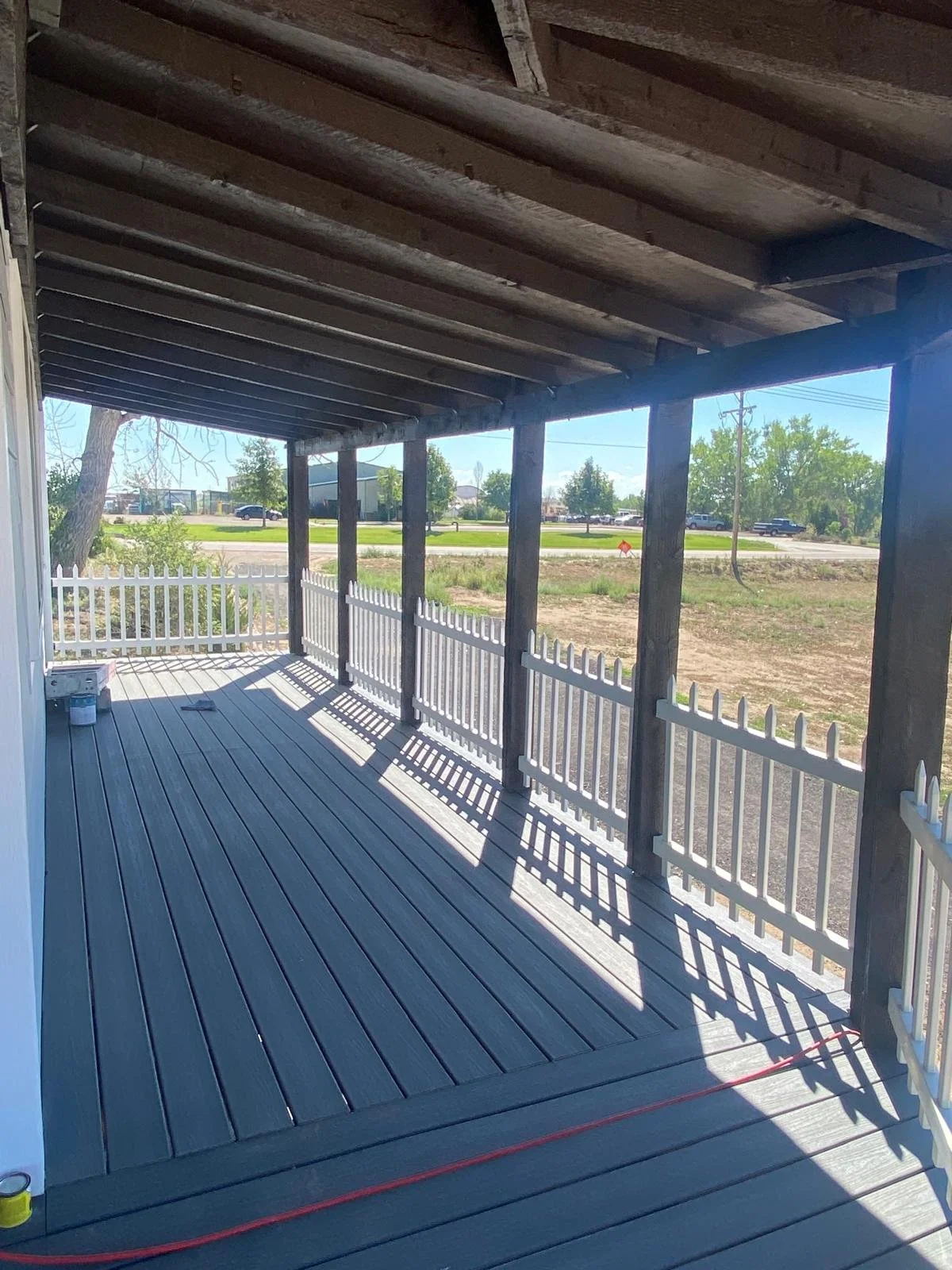 View of a wooden porch with a white picket fence railing, shadows cast on the floor, on a sunny day, with a grassy area and trees in the background.