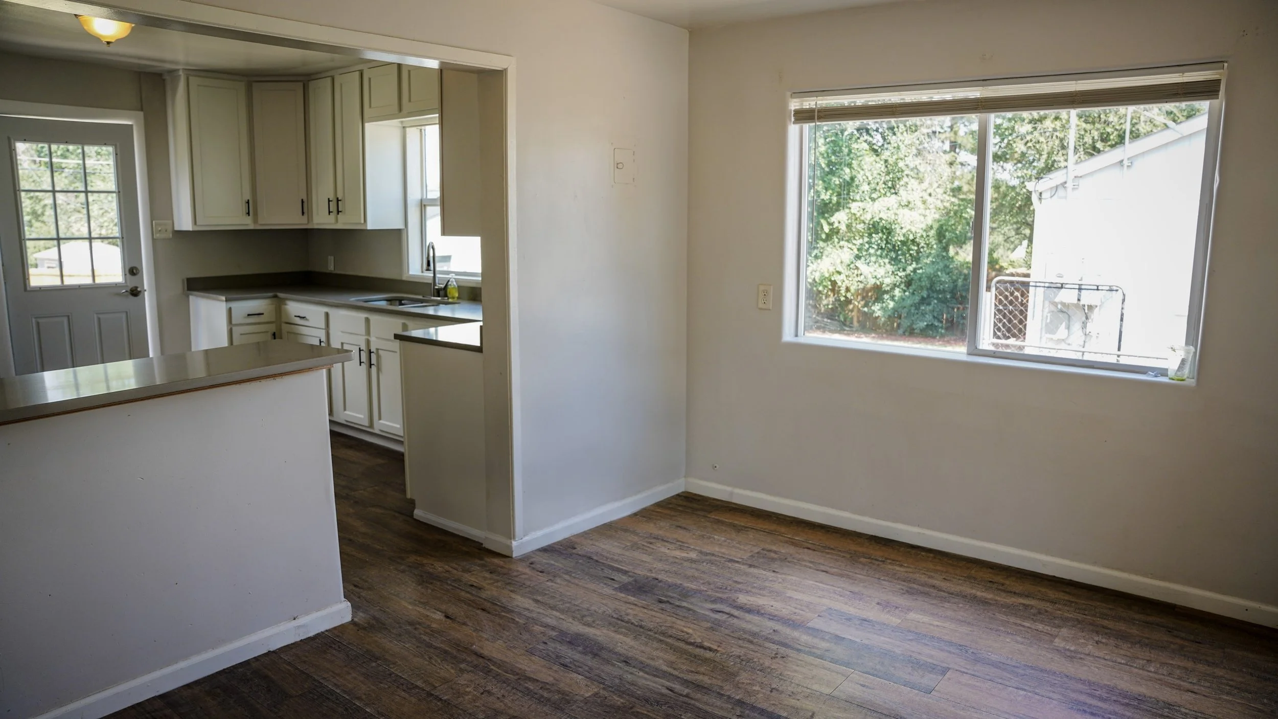 Empty dining area with large window overlooking backyard, adjacent to kitchen with white cabinets and a door leading outside.