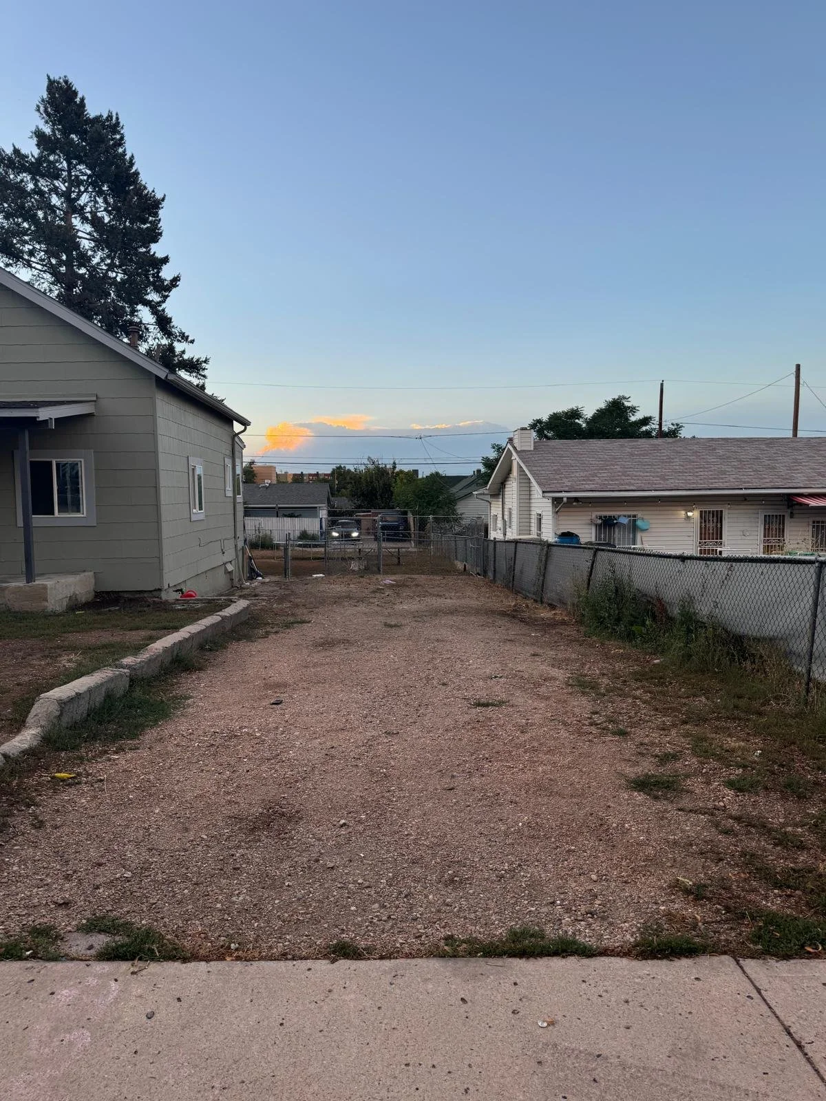 Empty gravel driveway between two houses, fence on the right and a small sidewalk in front, with trees and a sky with clouds in the background.