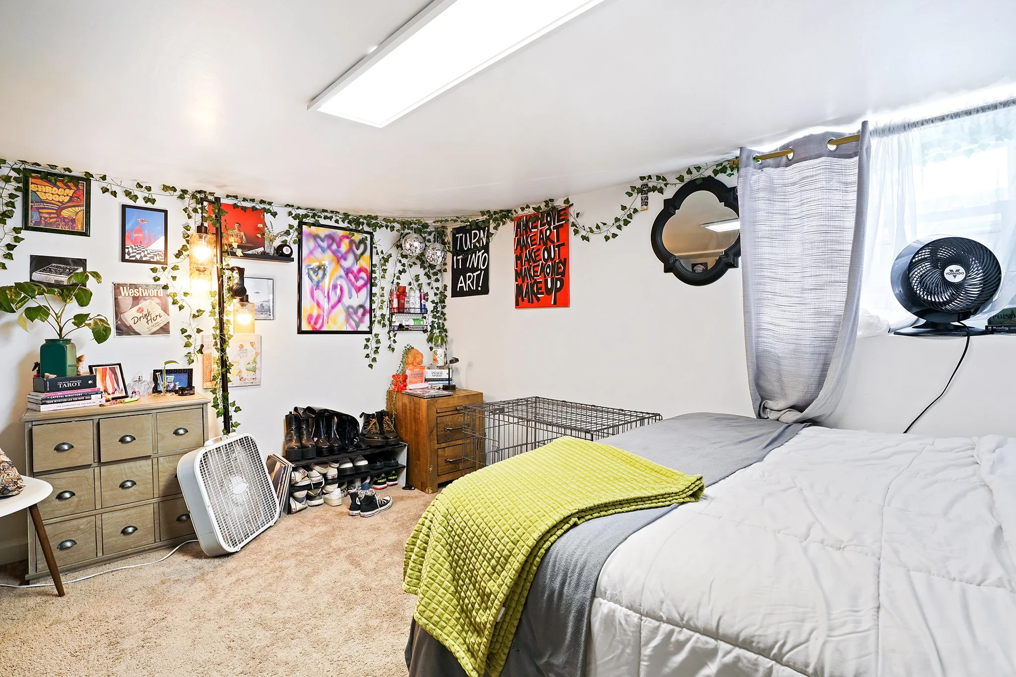 A messy bedroom with a bed, wall art, fairy lights, a dresser, a fan, shoes, and various decorative items.