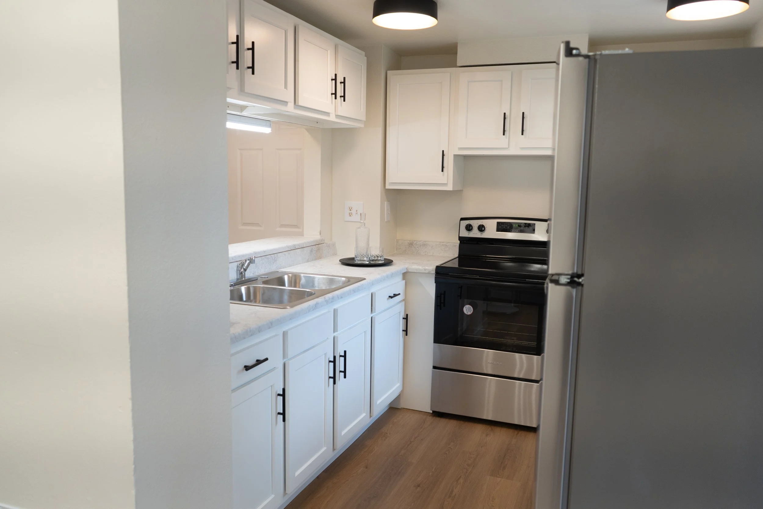 A compact kitchen with white cabinets, a stainless steel oven, a double sink, and a gray refrigerator. Wooden flooring and black cabinet handles complete the decor.