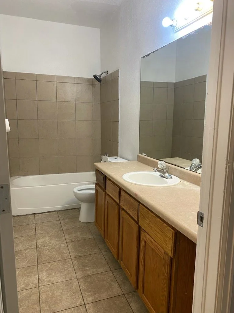 A small bathroom with a bathtub, toilet, and wooden vanity with a sink. A large mirror is above the vanity, and a row of lights is above the mirror. The walls have beige tiles in the shower area and are painted white elsewhere. The floor is tiled wit