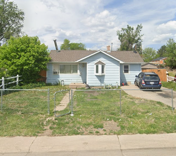 A light blue, single-story house with a small front yard, a black car parked on the driveway, and a chain-link fence around the yard.
