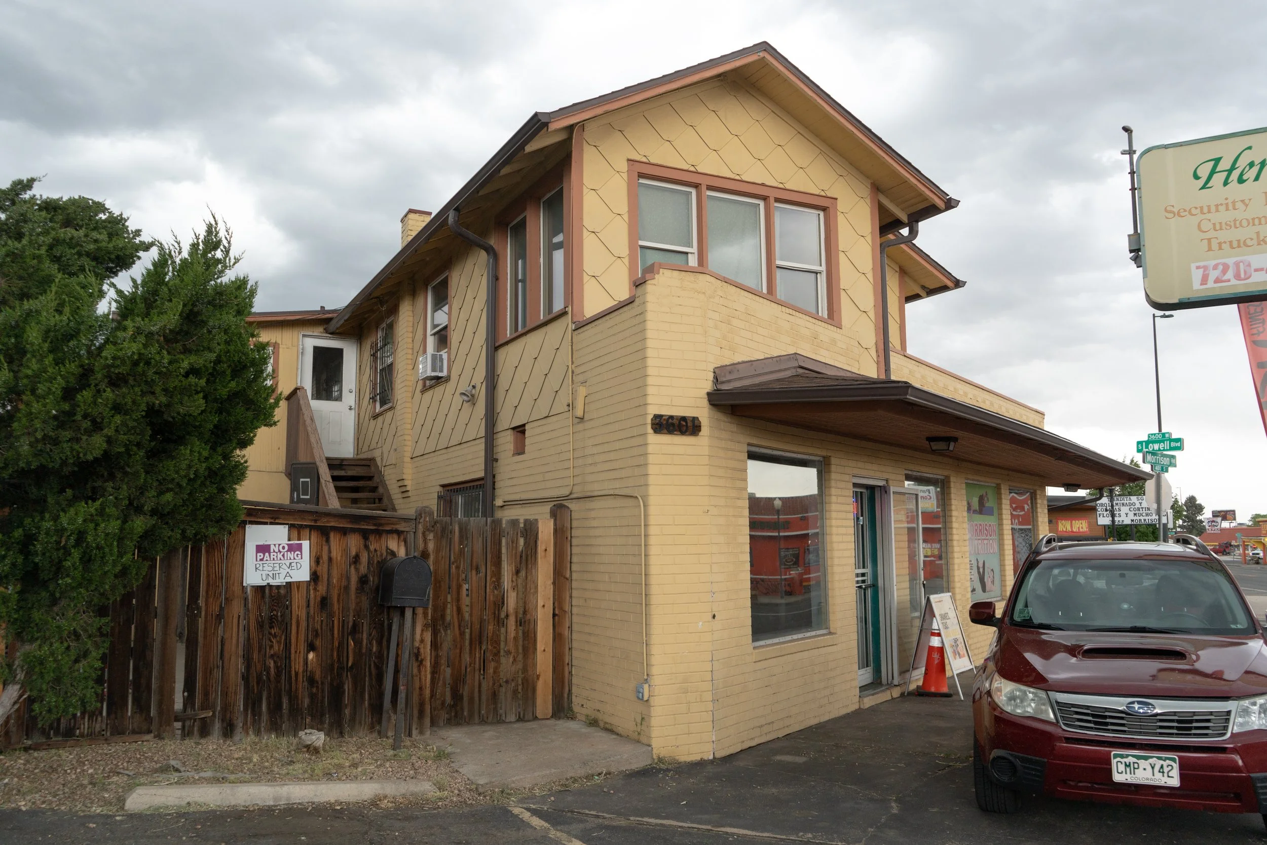 A two-story yellow brick building with windows, a small balcony, and a wooden staircase at the side, adjacent to a parking lot with a maroon Subaru car in front, and a wooden fence with a 'No Parking Reserved' sign. Overcast sky.