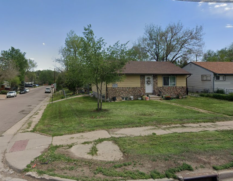 A small house with a stone and yellow exterior, front yard with grass and a tree, sidewalk, and neighboring houses on a residential street.