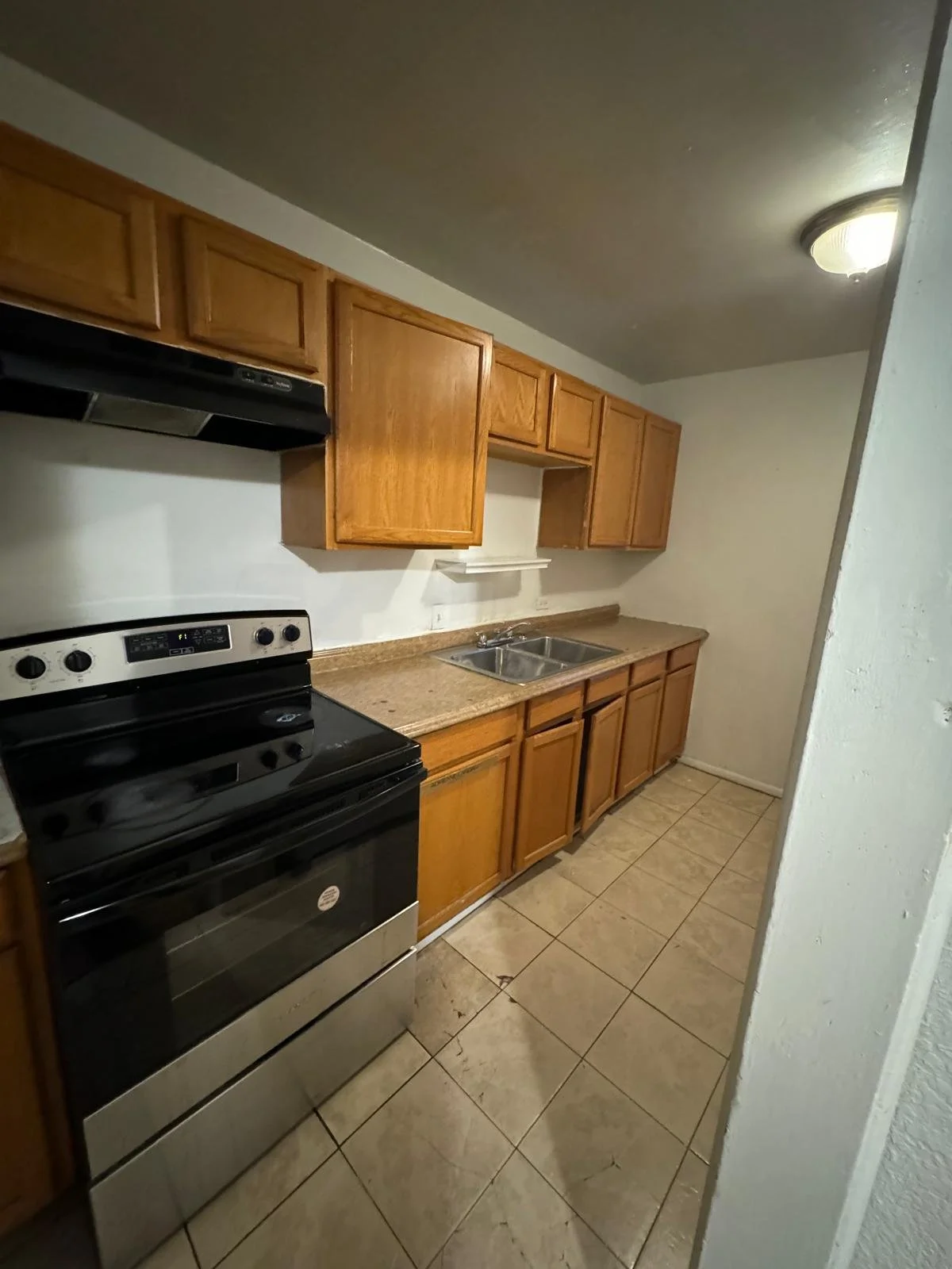 A kitchen with wooden cabinets, a black and stainless steel stove, double sink, beige tiled floor, and a white wall with a small shelf.